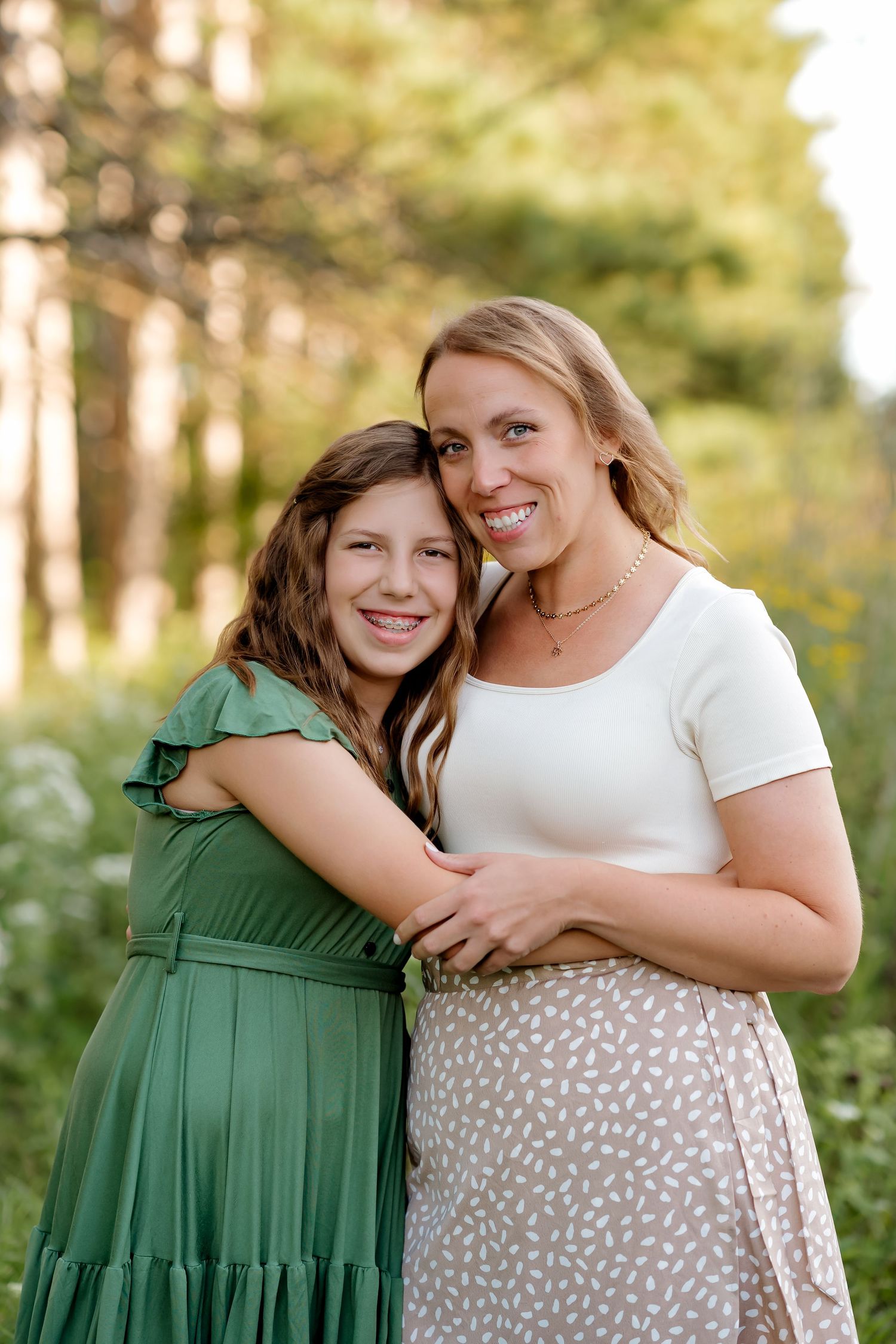 A smiling woman and young girl in green dress embrace outdoors among trees in a warm, sunlit portrait.