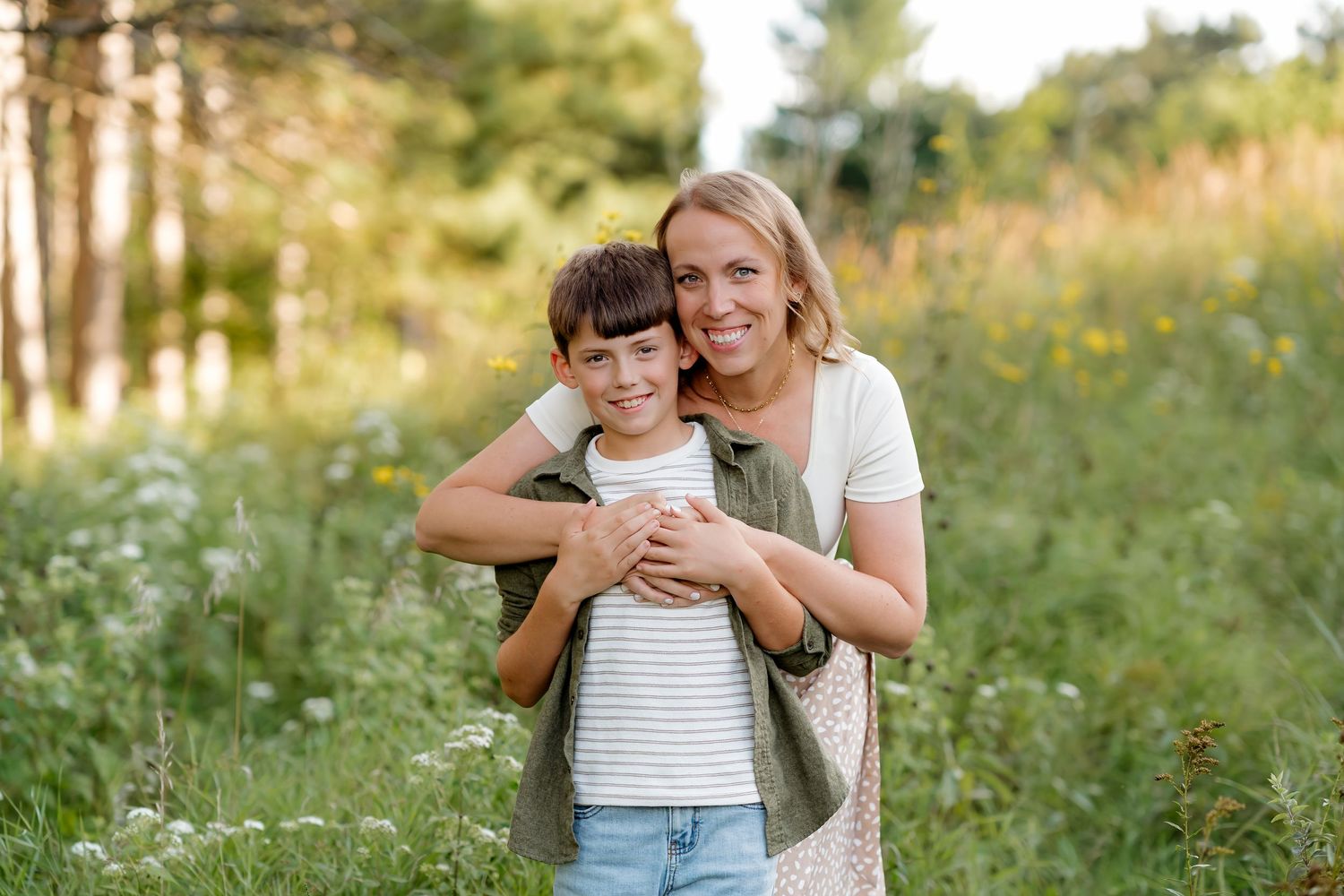 A woman and young boy embrace and smile in a sunlit meadow with wildflowers and trees in the background.