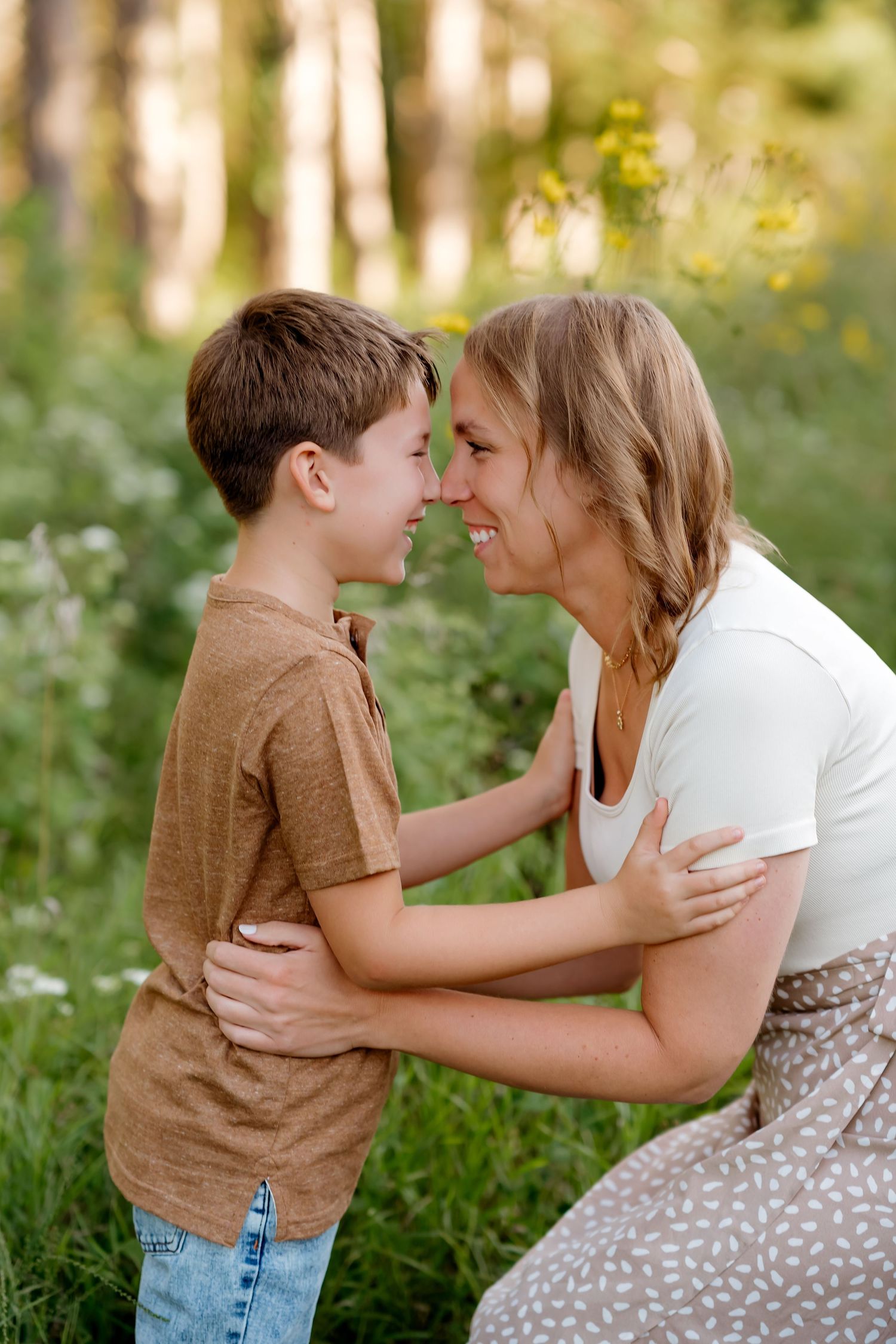 A mother and young boy share a joyful nose-to-nose moment outdoors, embracing in a sunny green meadow.