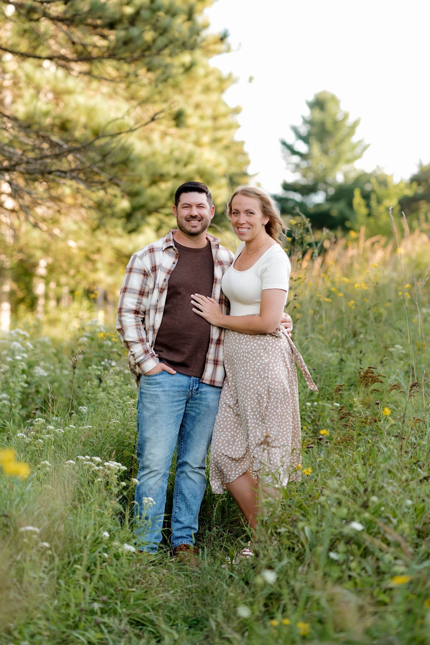 A smiling couple poses together in a lush green meadow surrounded by wildflowers and pine trees during golden hour.