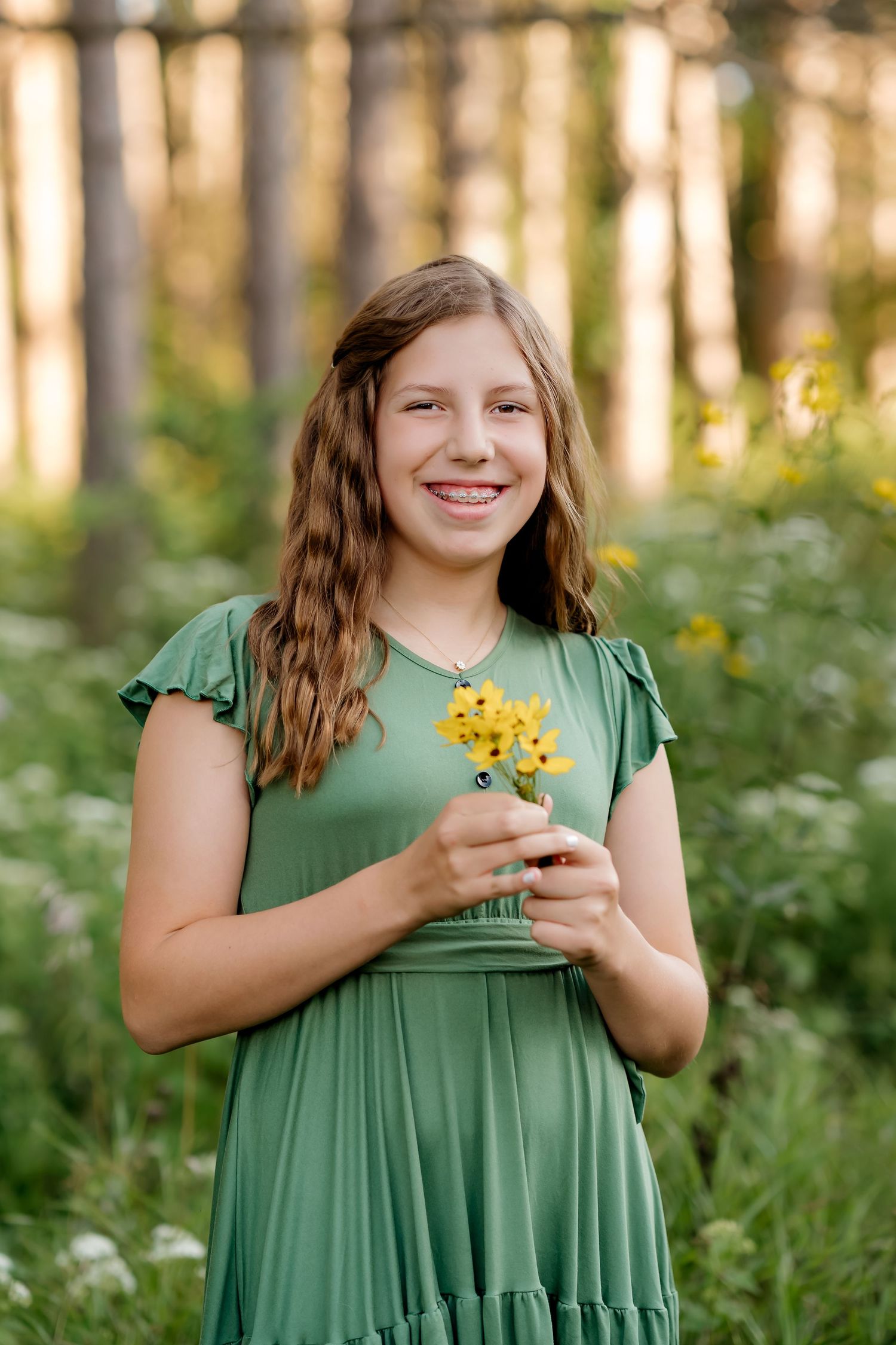 Smiling girl in green dress holding yellow wildflowers in a lush forest setting.