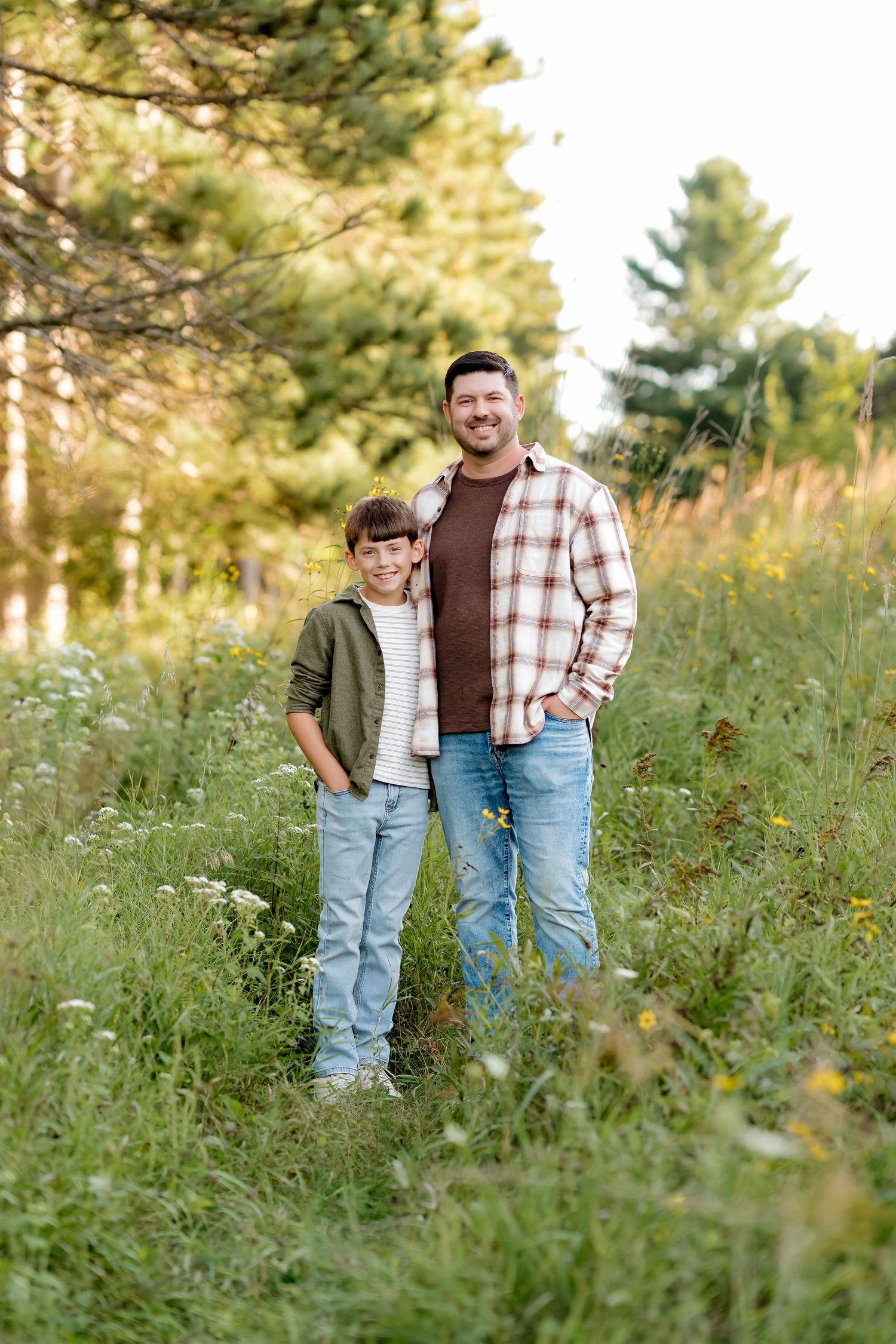 A smiling father and son stand together in a lush green meadow with wildflowers and trees in the background.