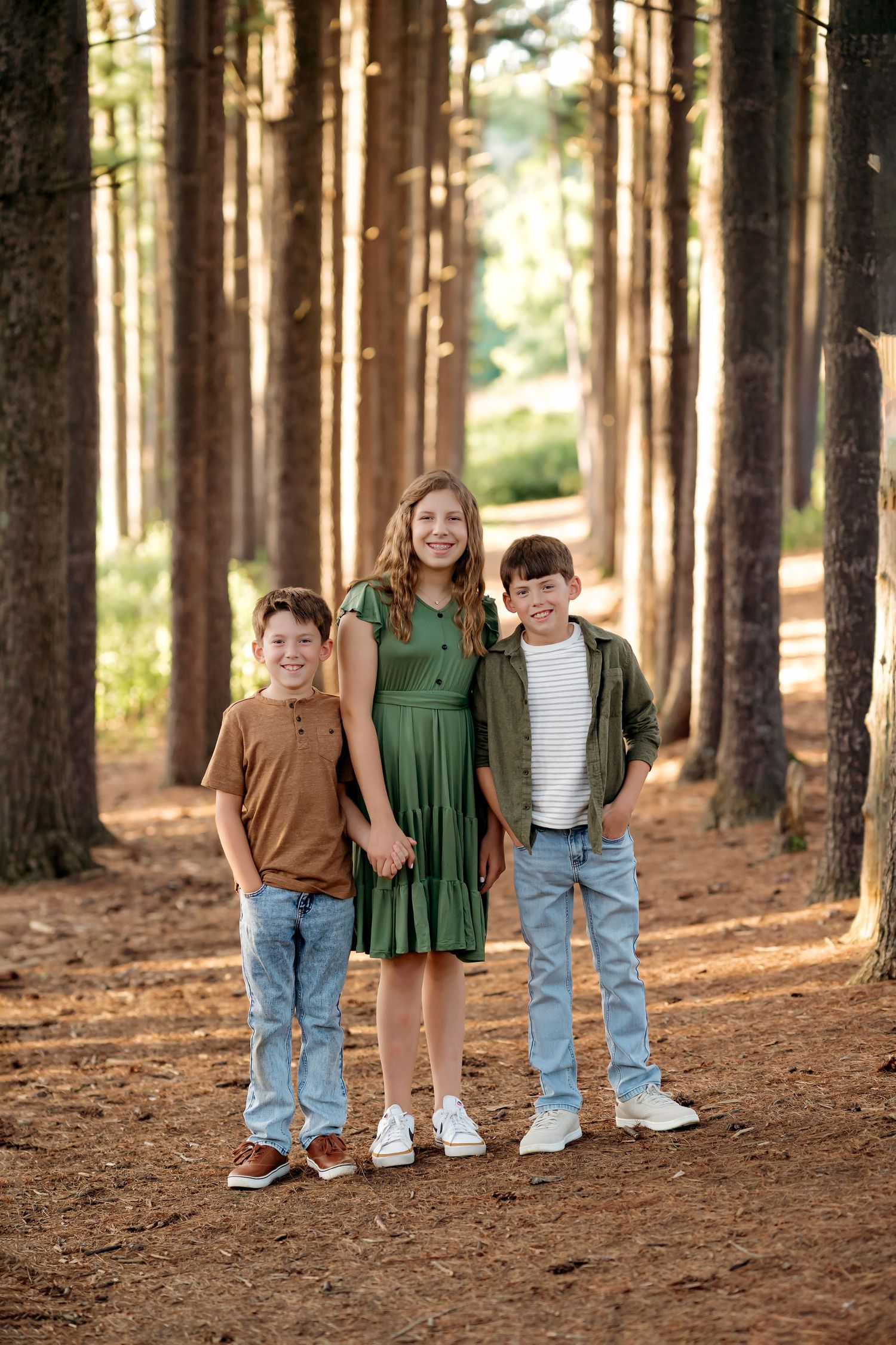 Three smiling children pose among tall pine trees on a forest path, wearing earth tones and denim.