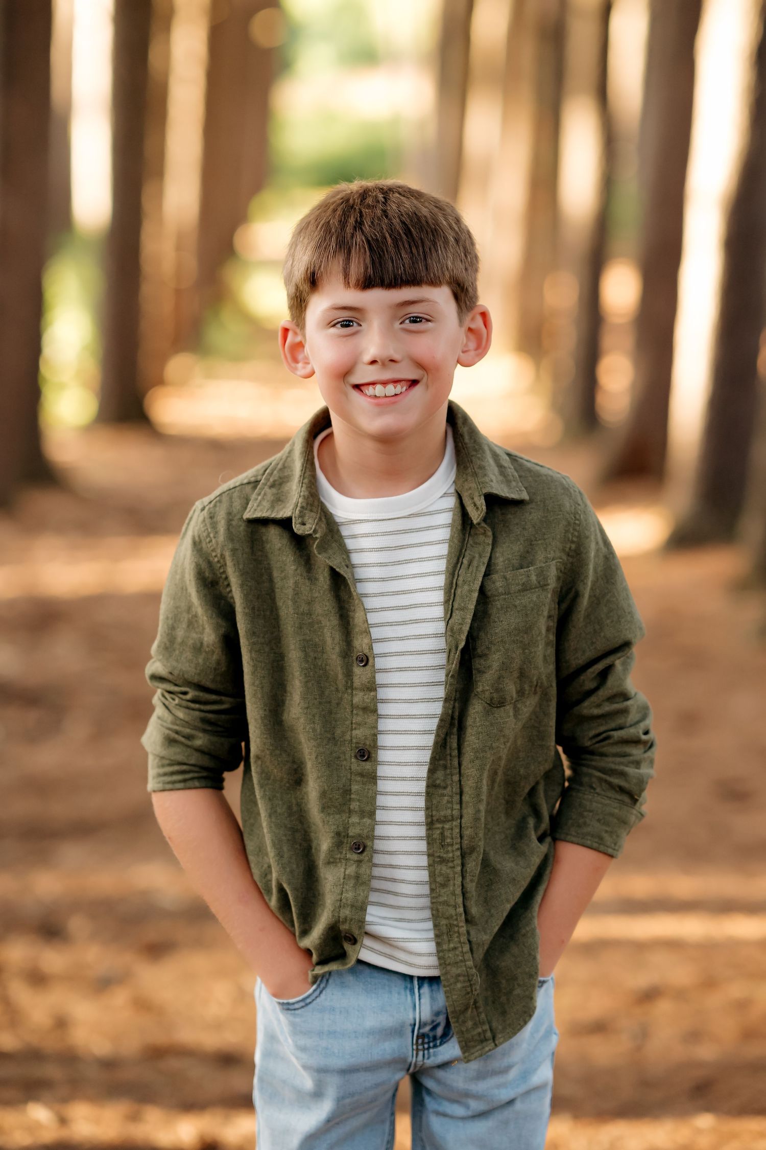 Smiling boy in green shirt and jeans stands in a tree-lined path outdoors.