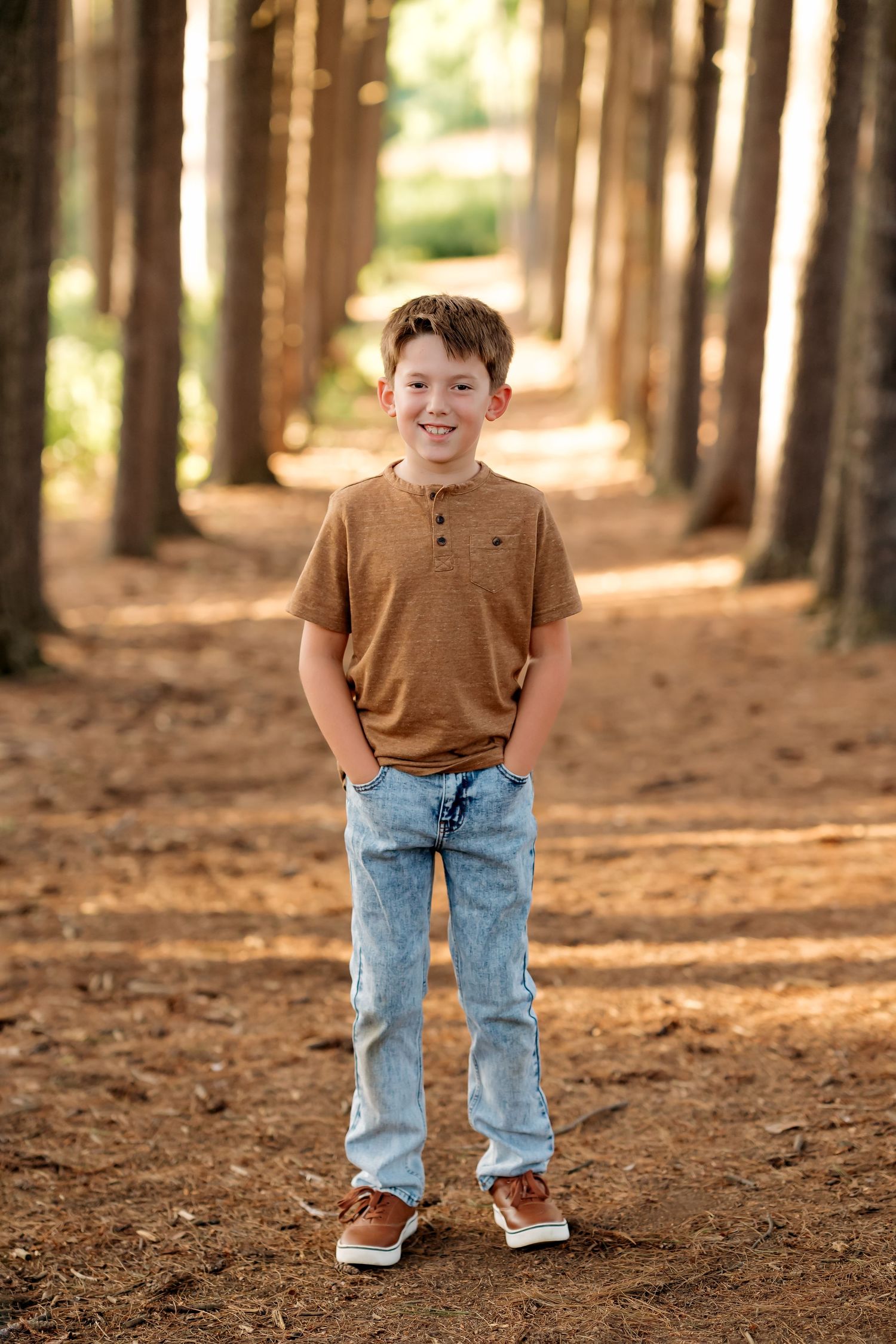 A smiling boy in a brown shirt and jeans stands on a pine tree-lined path with hands in pockets.