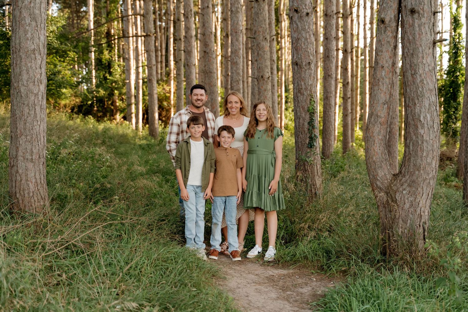 Family of five smiling together on a forest path surrounded by tall pine trees and lush green grass in warm sunlight.