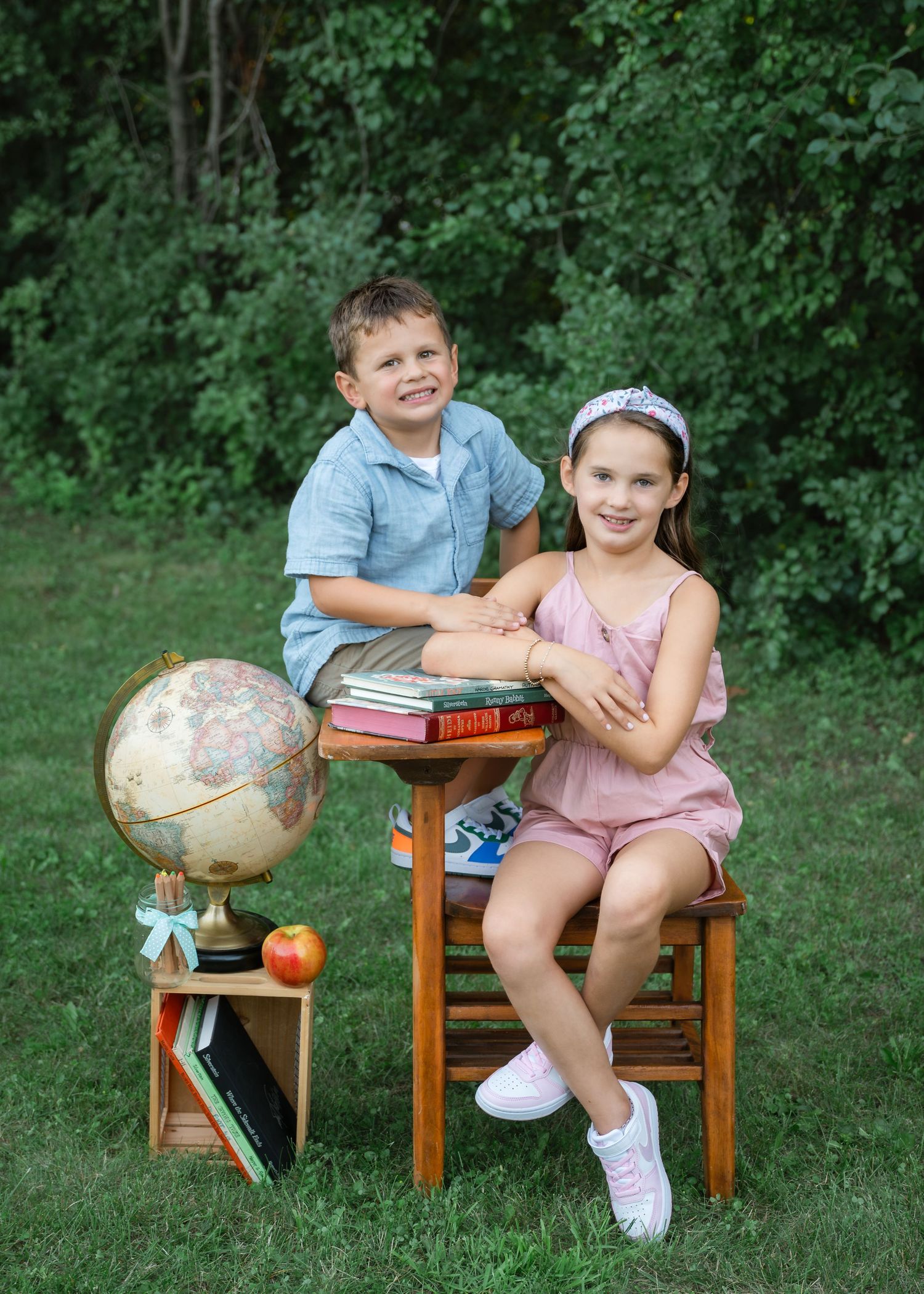 Two children pose outdoors with a globe, apple, and books for a back-to-school portrait on green grass.