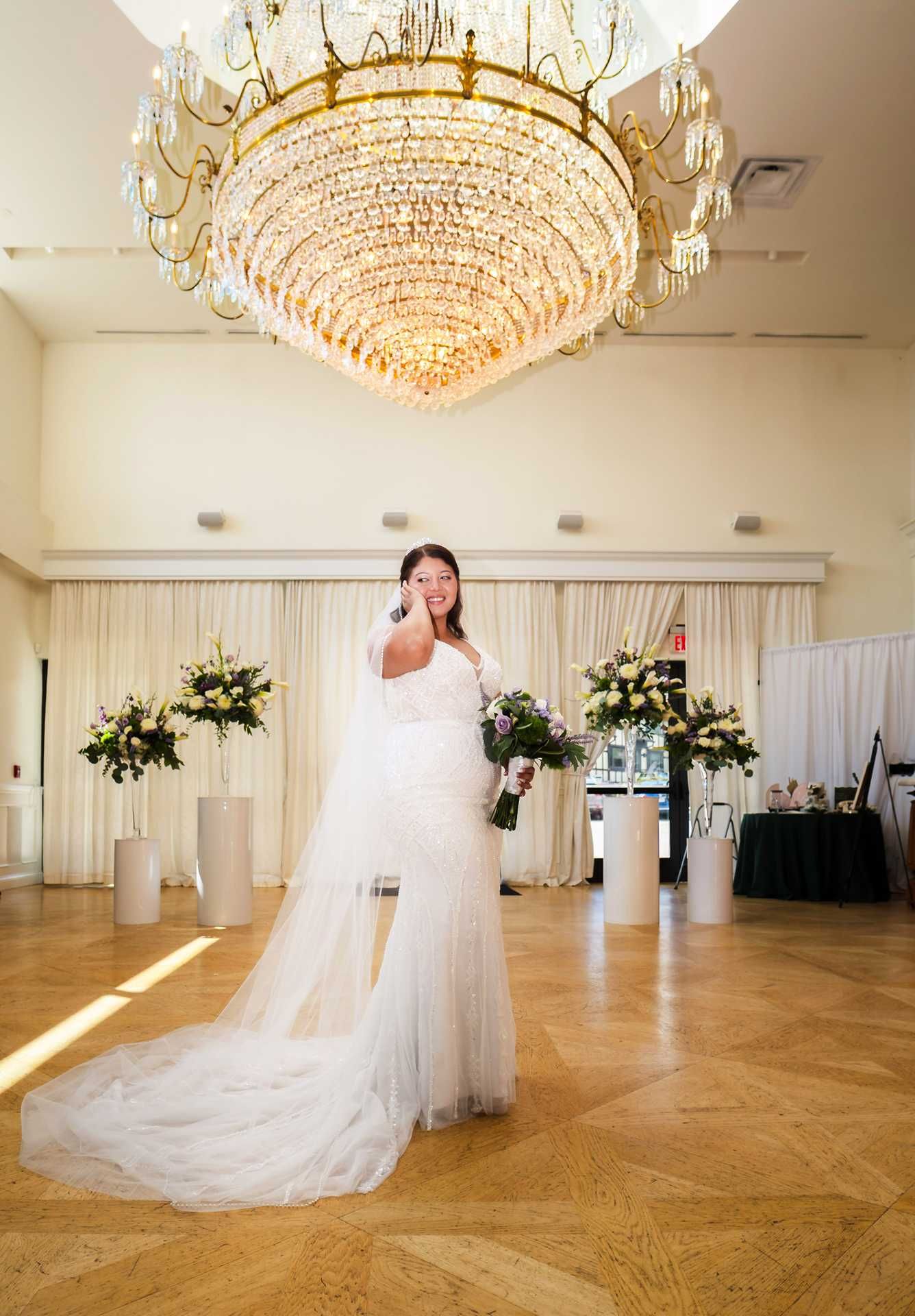 Bride in white mermaid gown with long veil stands under grand crystal chandelier in elegant ballroom at the view wedding venue