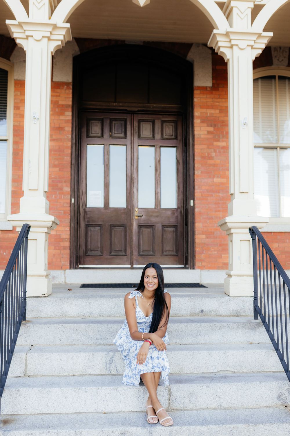 Young woman in floral dress sitting on steps of elegant brick building with ornate white columns and wooden doors.