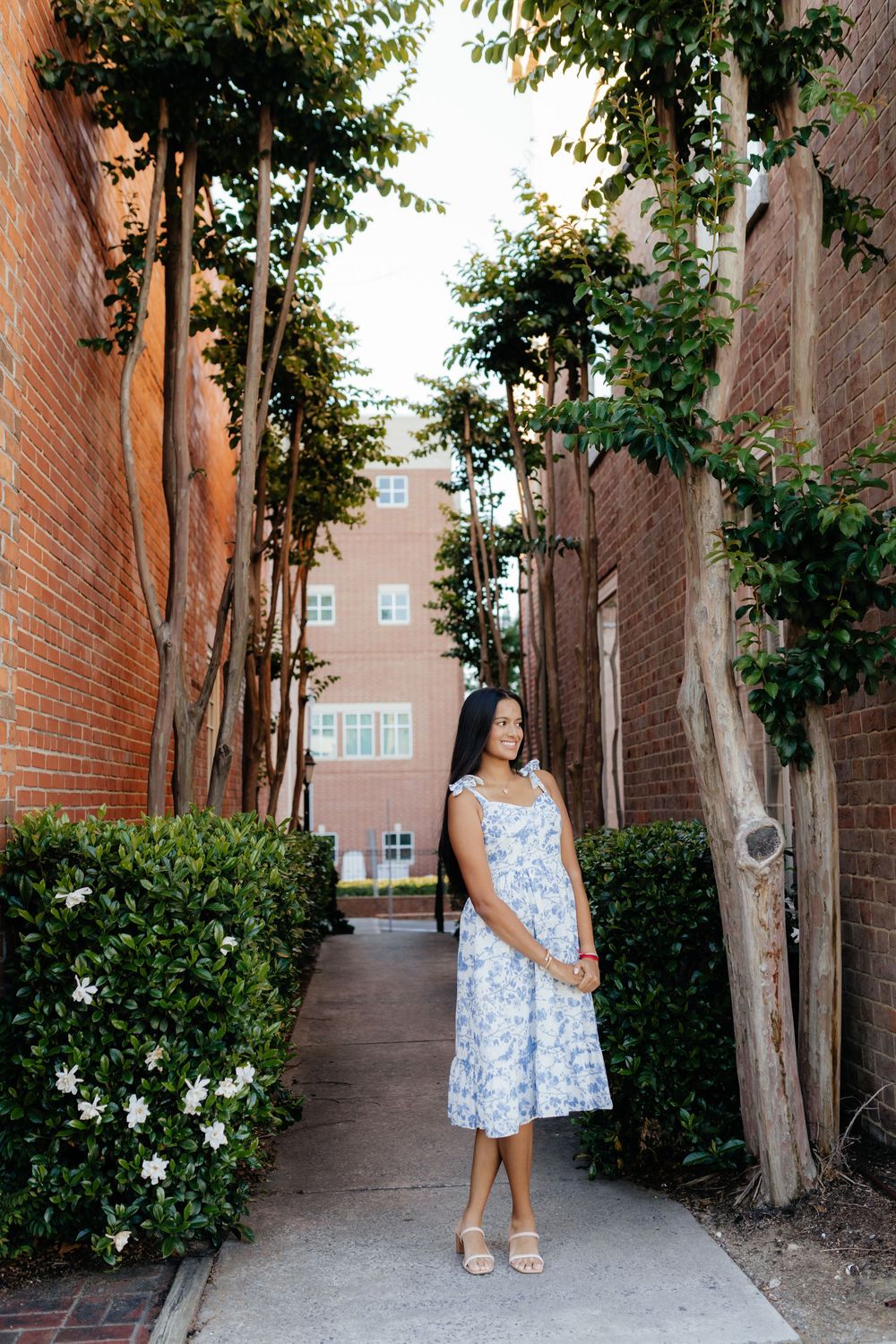 Woman in blue floral sundress standing in a charming brick alleyway with lush greenery and white flowers.