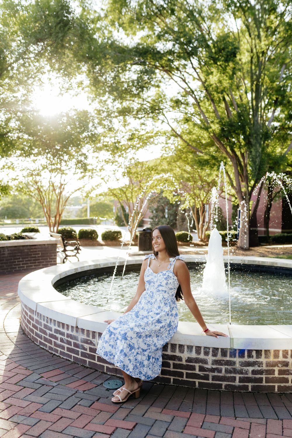 Woman in blue floral maxi dress sitting on brick fountain in sunny park with lush green trees.
