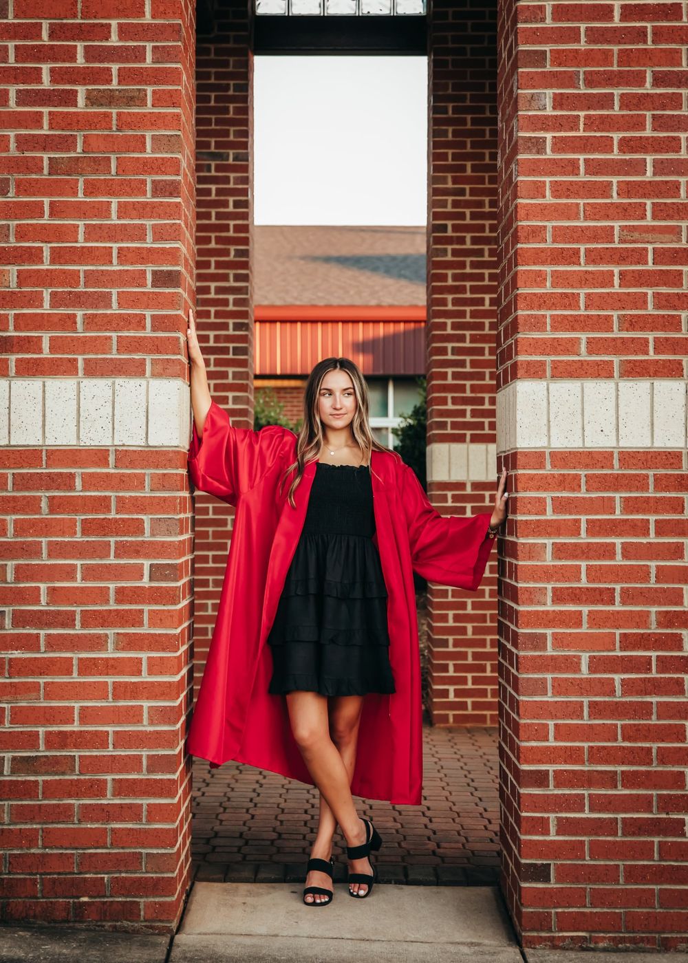 Young woman in red graduation gown and black dress poses in brick doorway for senior portrait photo.