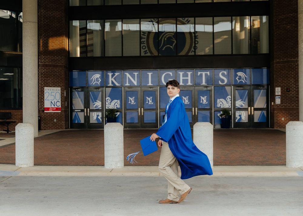A graduate in a blue cap and gown poses proudly in front of a Knights school entrance with trophy cases.