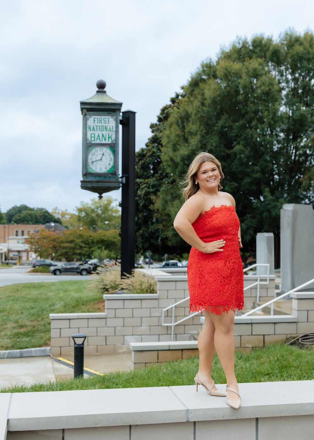 Woman in red lace dress posing near First National Bank clock in a small town setting.