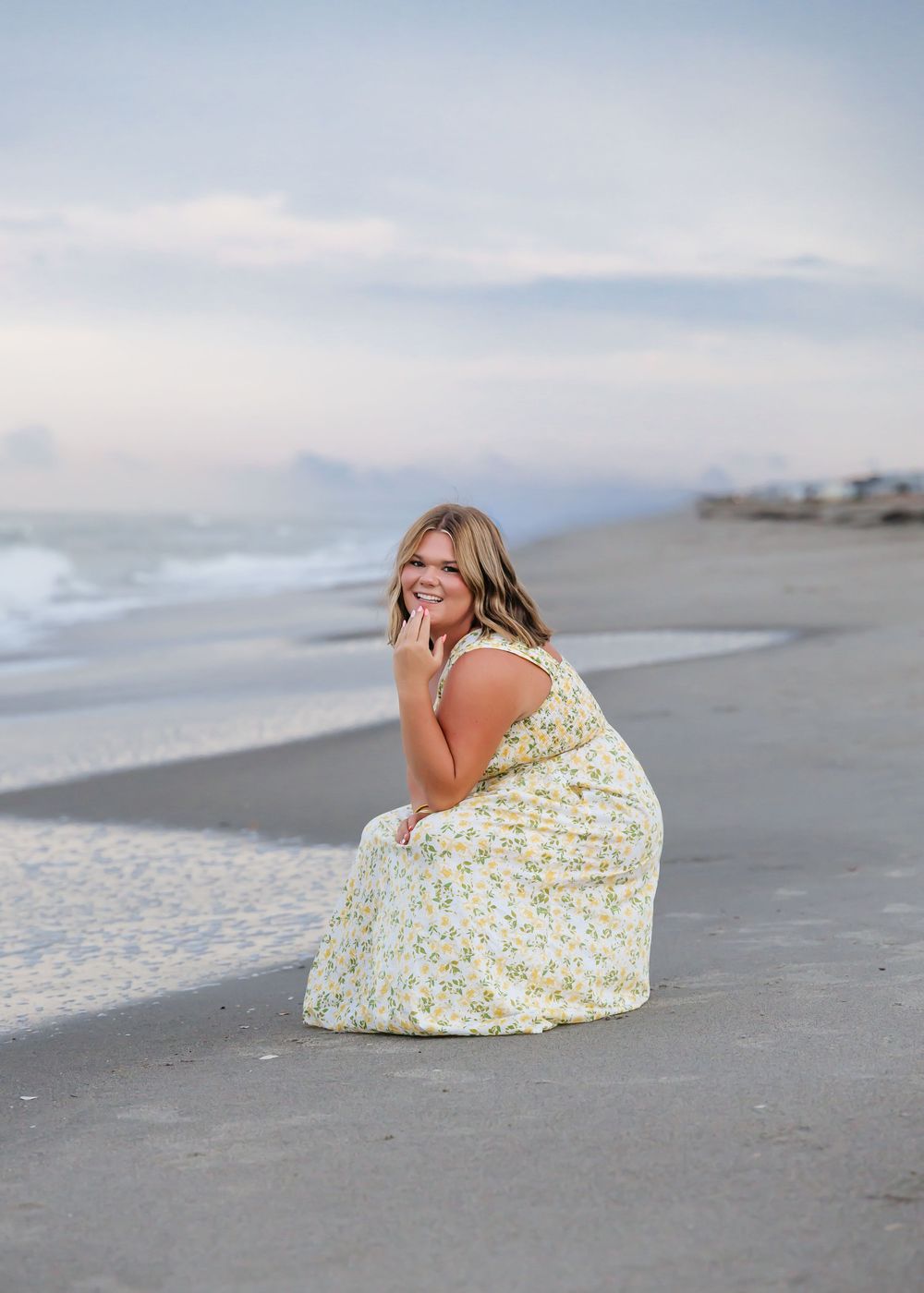 A smiling woman in a floral maxi dress sits on a sandy beach at dusk, with gentle waves and pastel sky behind her.