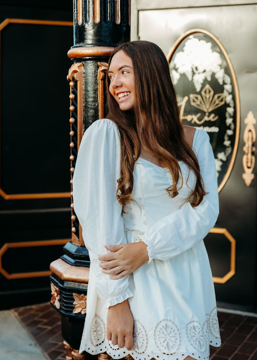 Young woman in white eyelet dress with long sleeves smiles outdoors near ornate black and gold decor.
