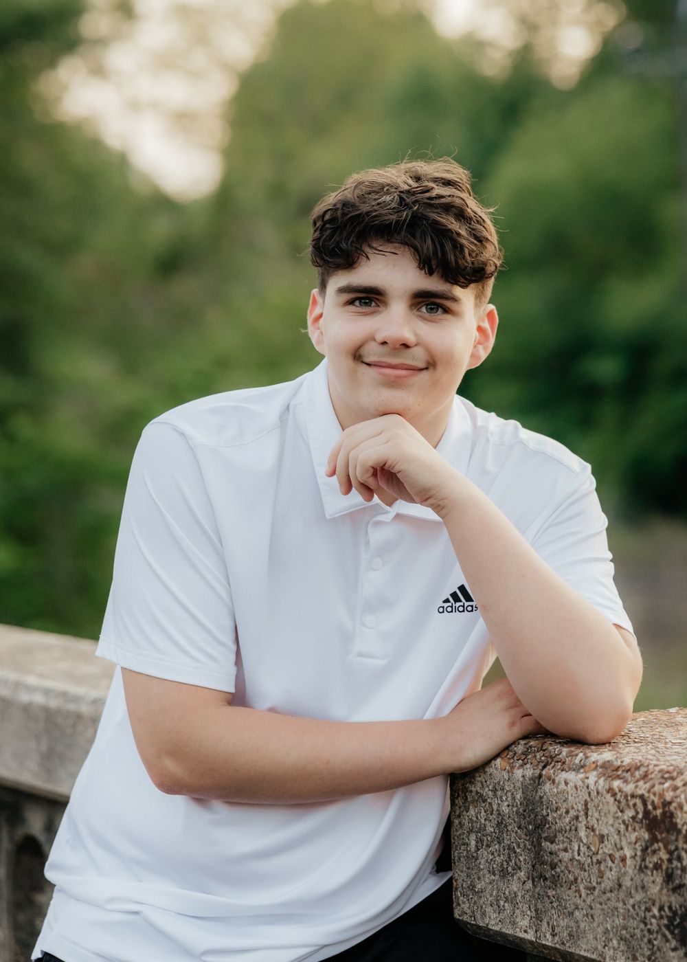 Young man in white Adidas polo shirt leaning on stone wall outdoors with green trees in background.