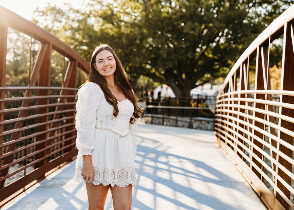 Young woman in white lace dress smiling on a bridge surrounded by trees in golden sunlight.