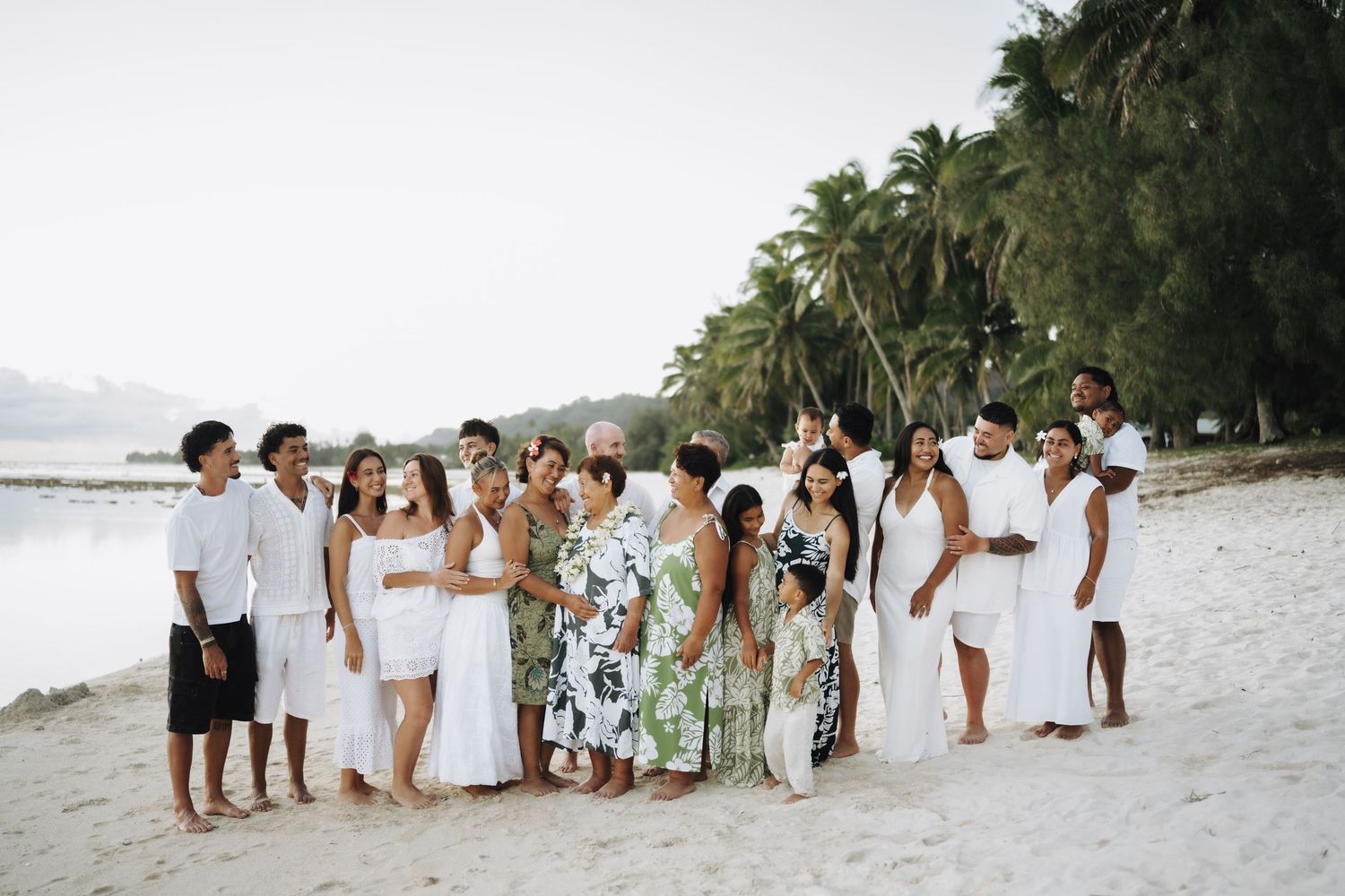 Large wedding party dressed in white gathered on a tropical beach surrounded by palm trees.
