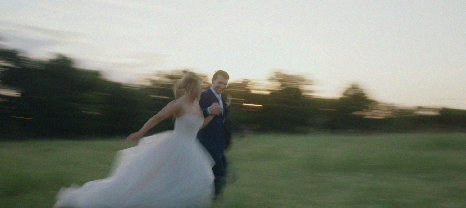 A bride and groom run joyfully through a lush green field at dusk in a romantic motion-blurred wedding photo.