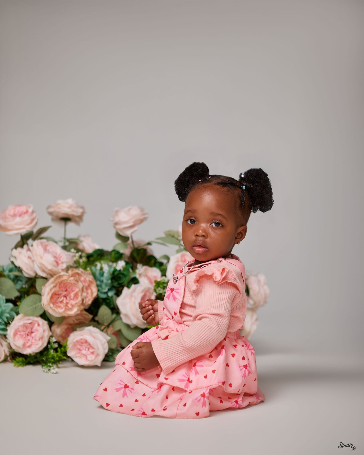 Adorable baby girl in pink dress with pigtails sitting beside blush roses in studio portrait.
