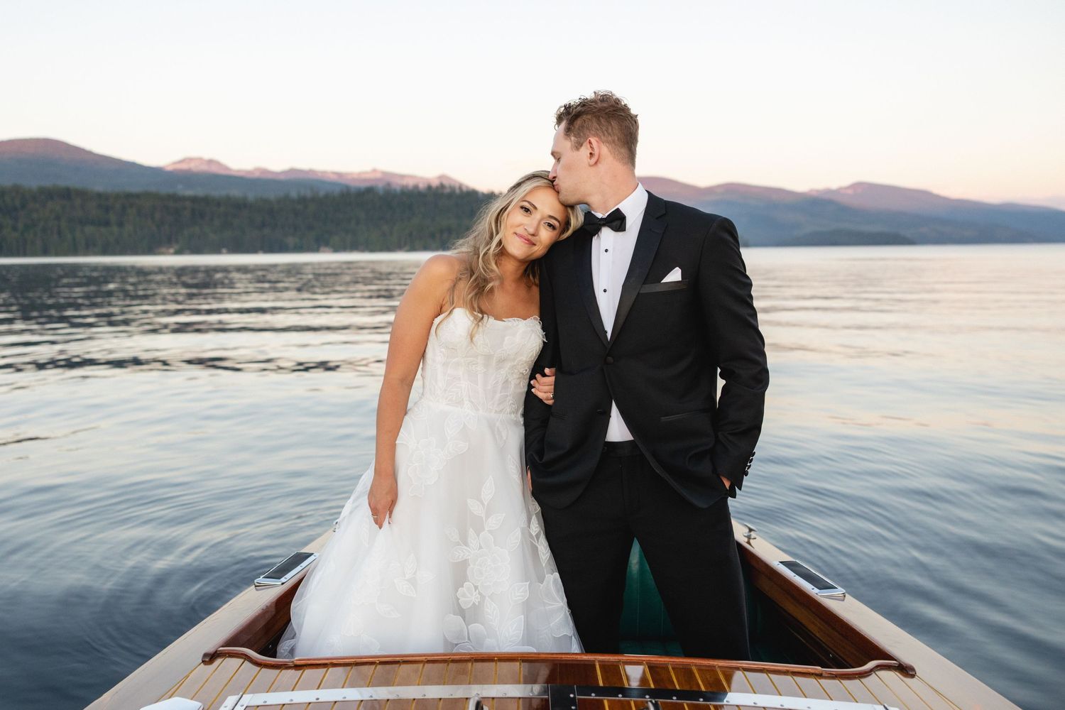 Bride and groom posing on a wooden boat at sunset with mountain lake backdrop in wedding attire.