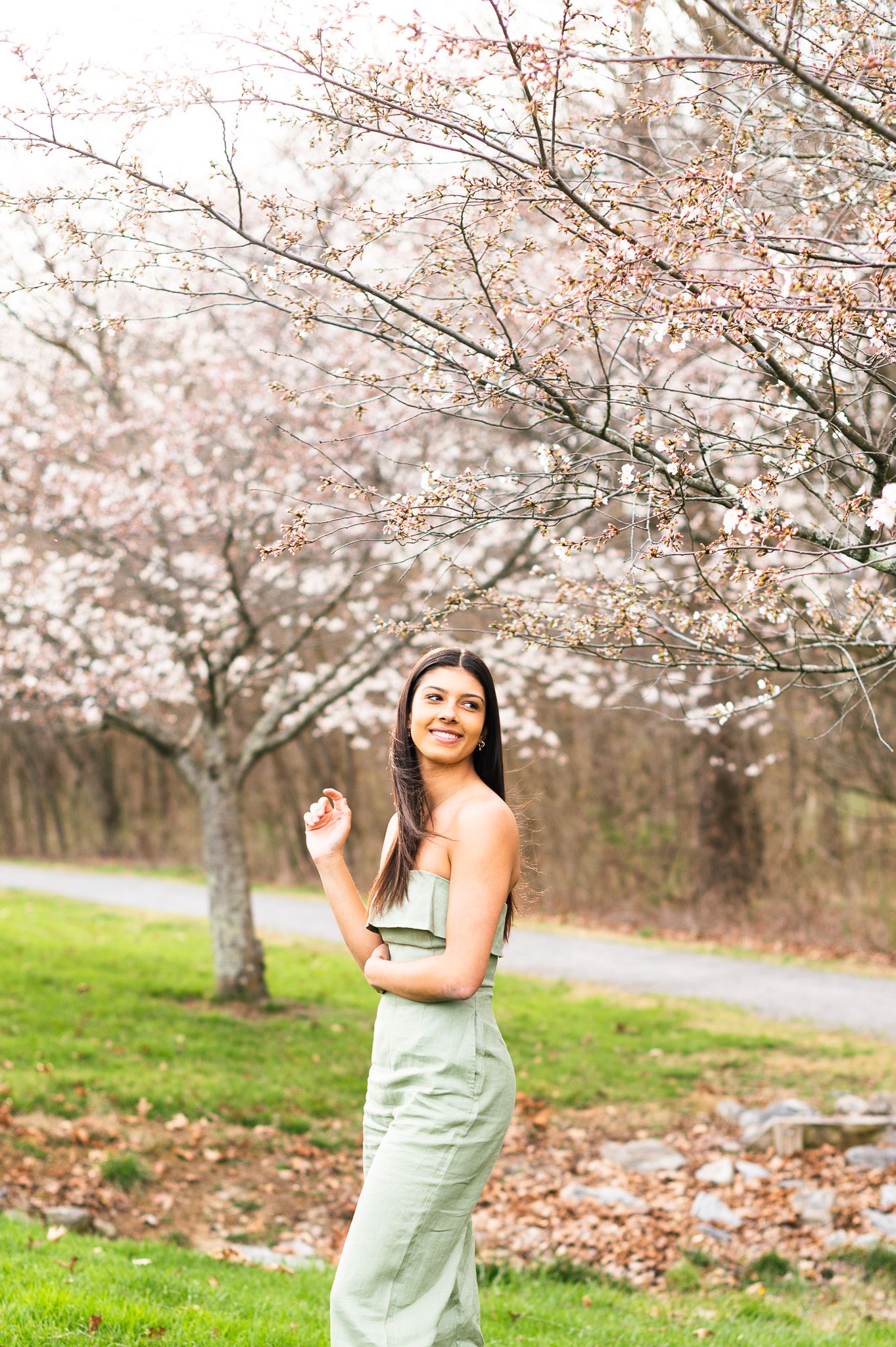 Young woman in sage green jumpsuit smiling under blooming cherry blossom trees on a lush green lawn.