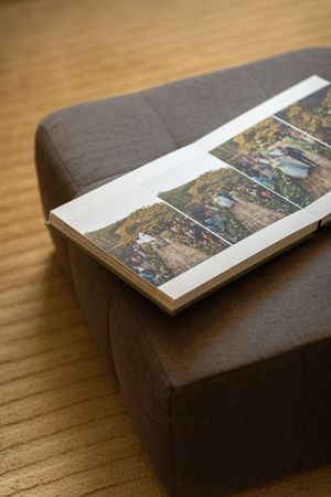 Open photo book displaying colorful garden images resting on a gray upholstered ottoman on a wooden floor.