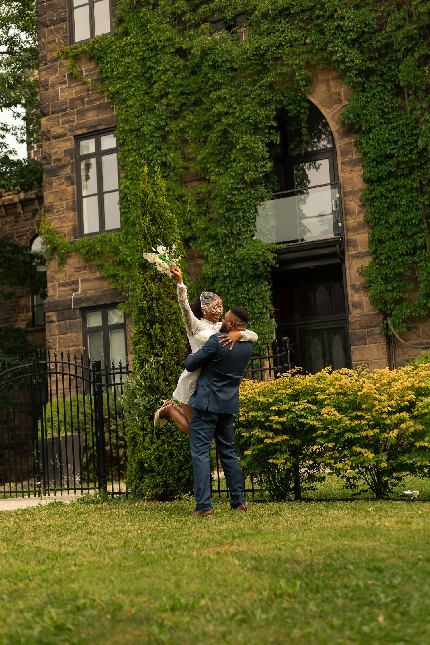 Groom lifting bride in white dress outside ivy-covered stone building, bride raising bouquet joyfully.