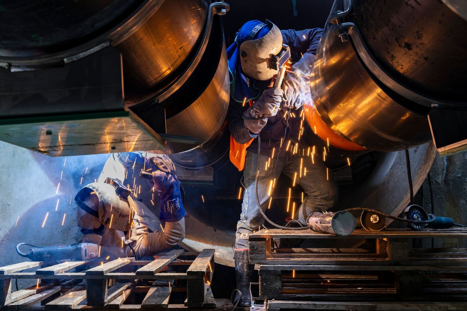 Two industrial workers grinding metal with sparks flying in a factory workshop setting.