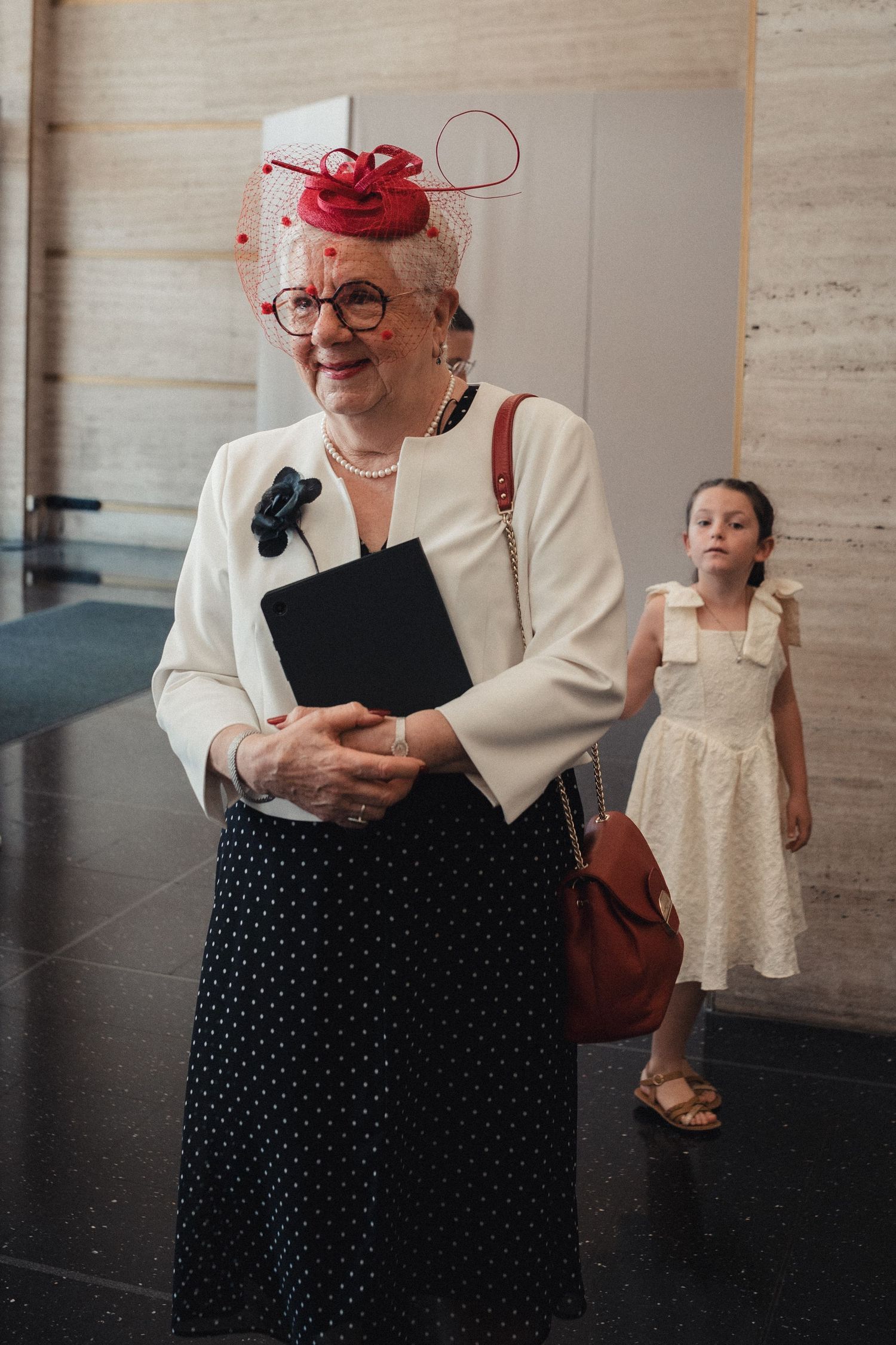 Elegant elderly woman in red fascinator hat and white jacket smiles, holding a black folder, with a young girl behind her.