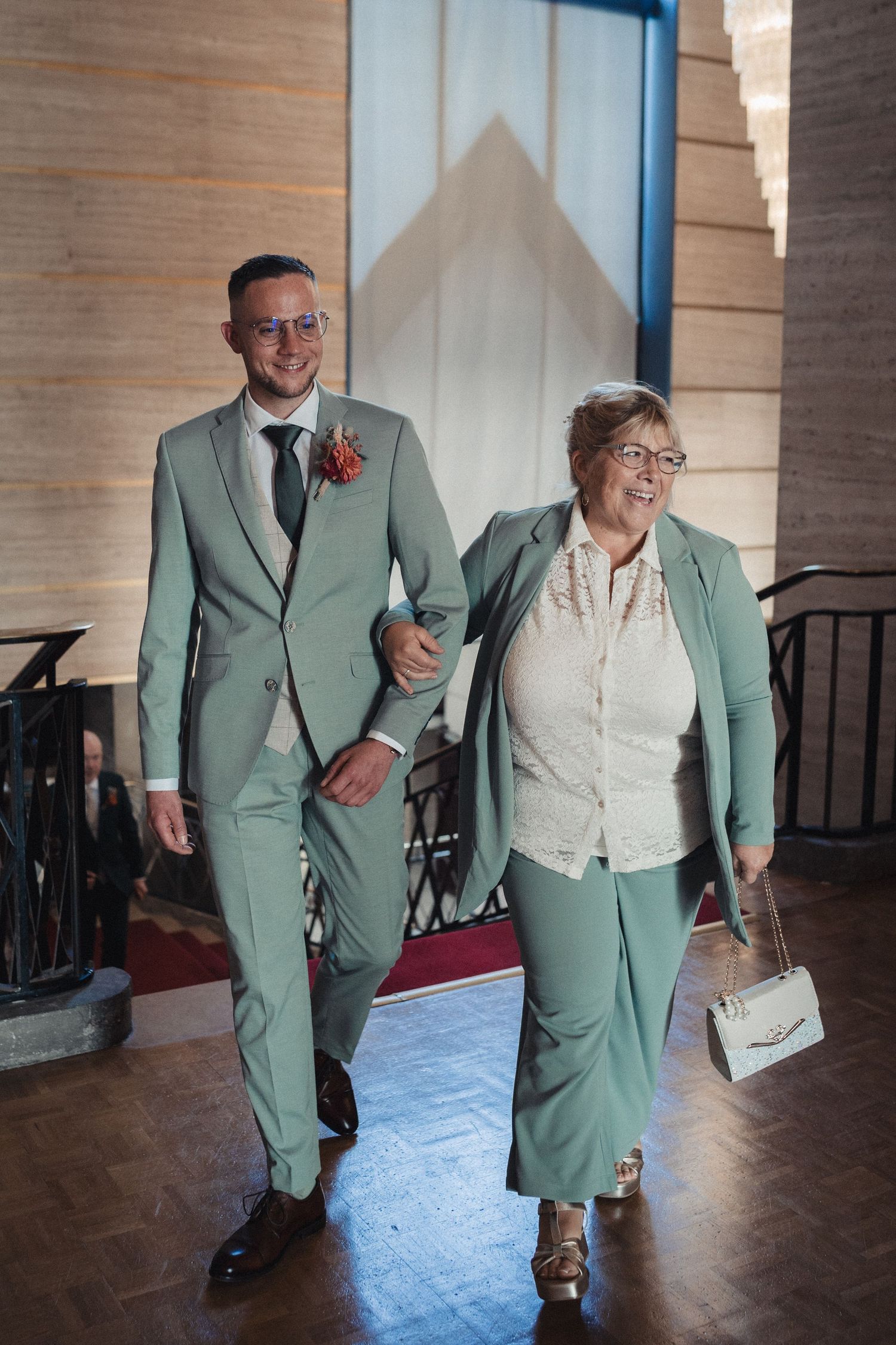 Groom in sage green suit walks arm-in-arm with elderly woman in matching green pantsuit at wedding venue.