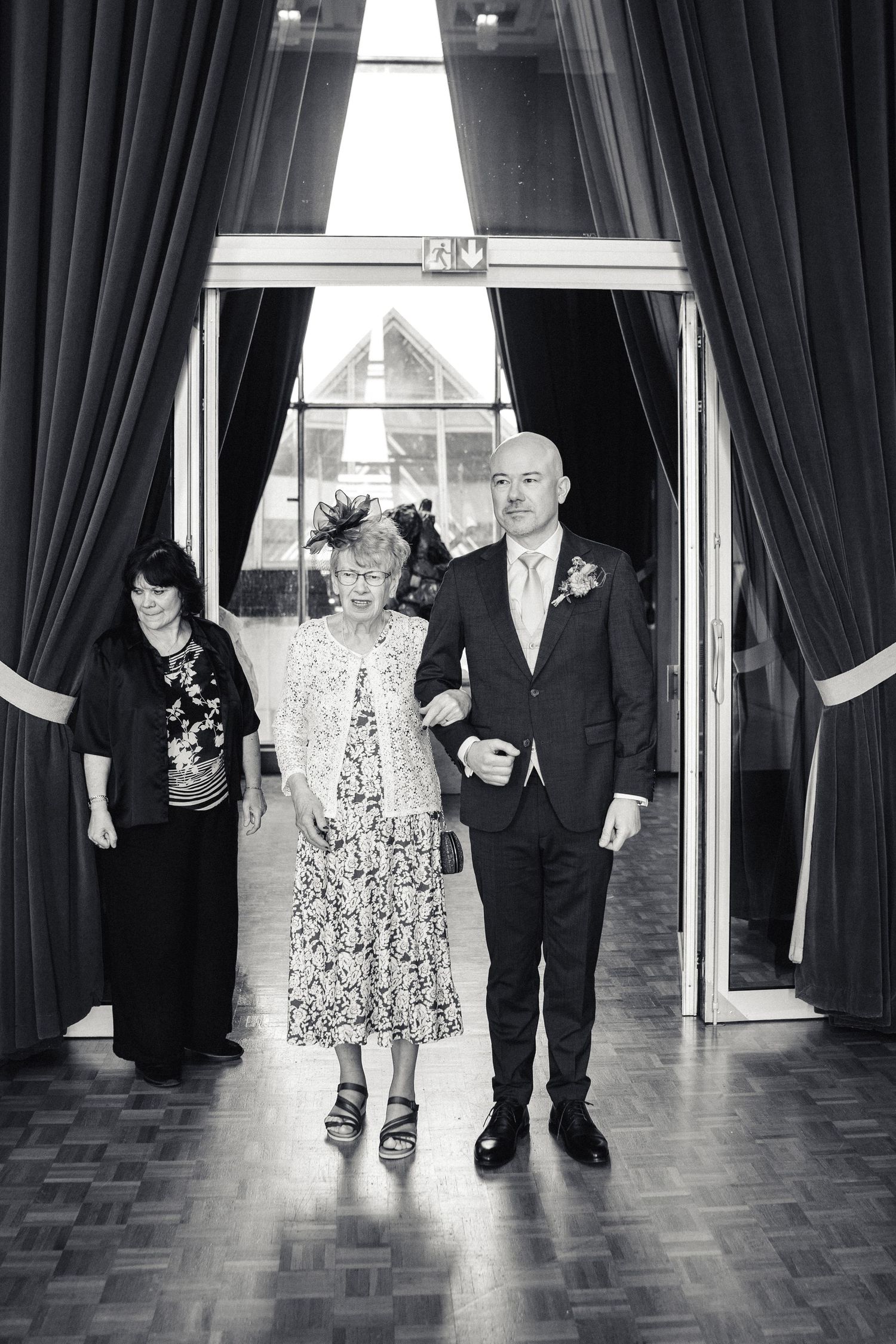 Black and white photo of groom escorting elderly woman into wedding venue ceremony.