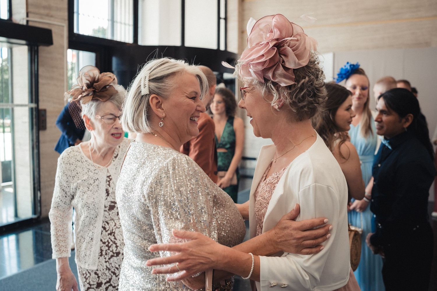 Two elegant women in formal attire warmly embrace at a wedding reception, wearing lace and floral accessories.