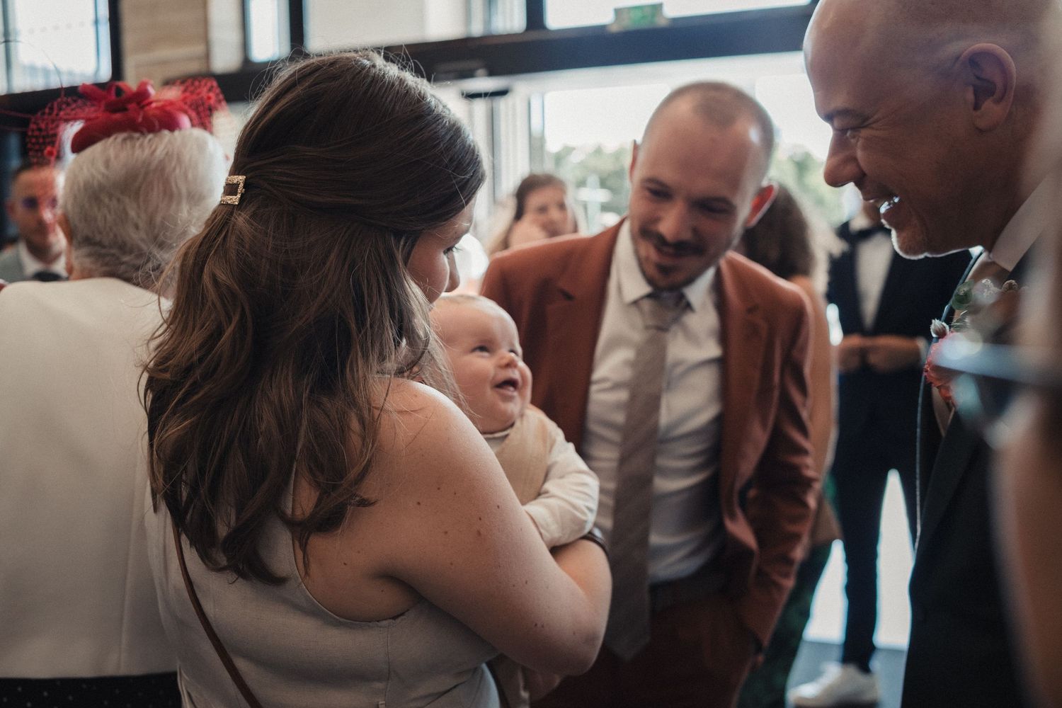 Adults gathered at a wedding reception admiring a baby held by a woman in a white dress.