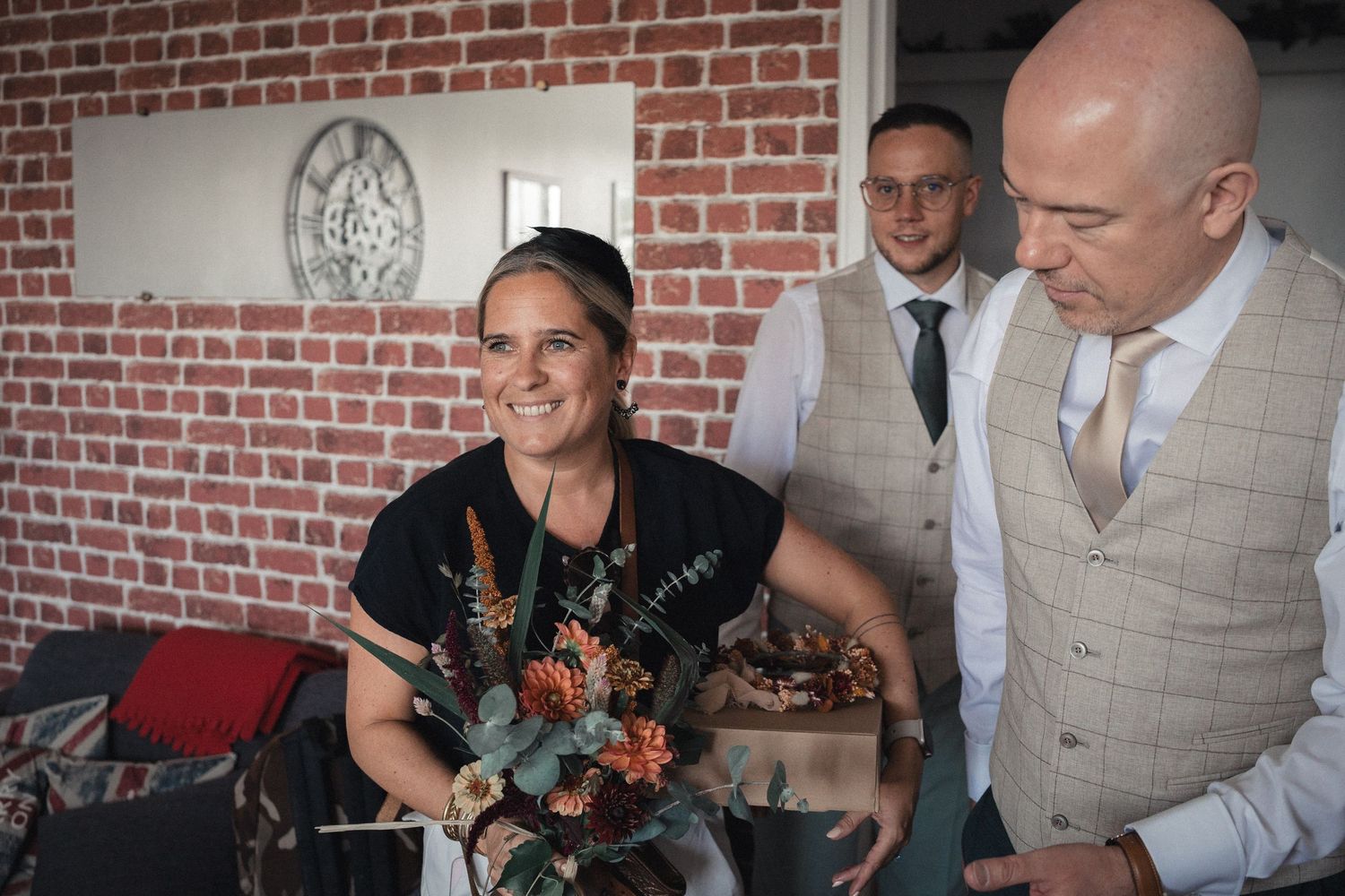 Woman holding a floral bouquet smiles at a formal event, with two men in suits standing nearby indoors.