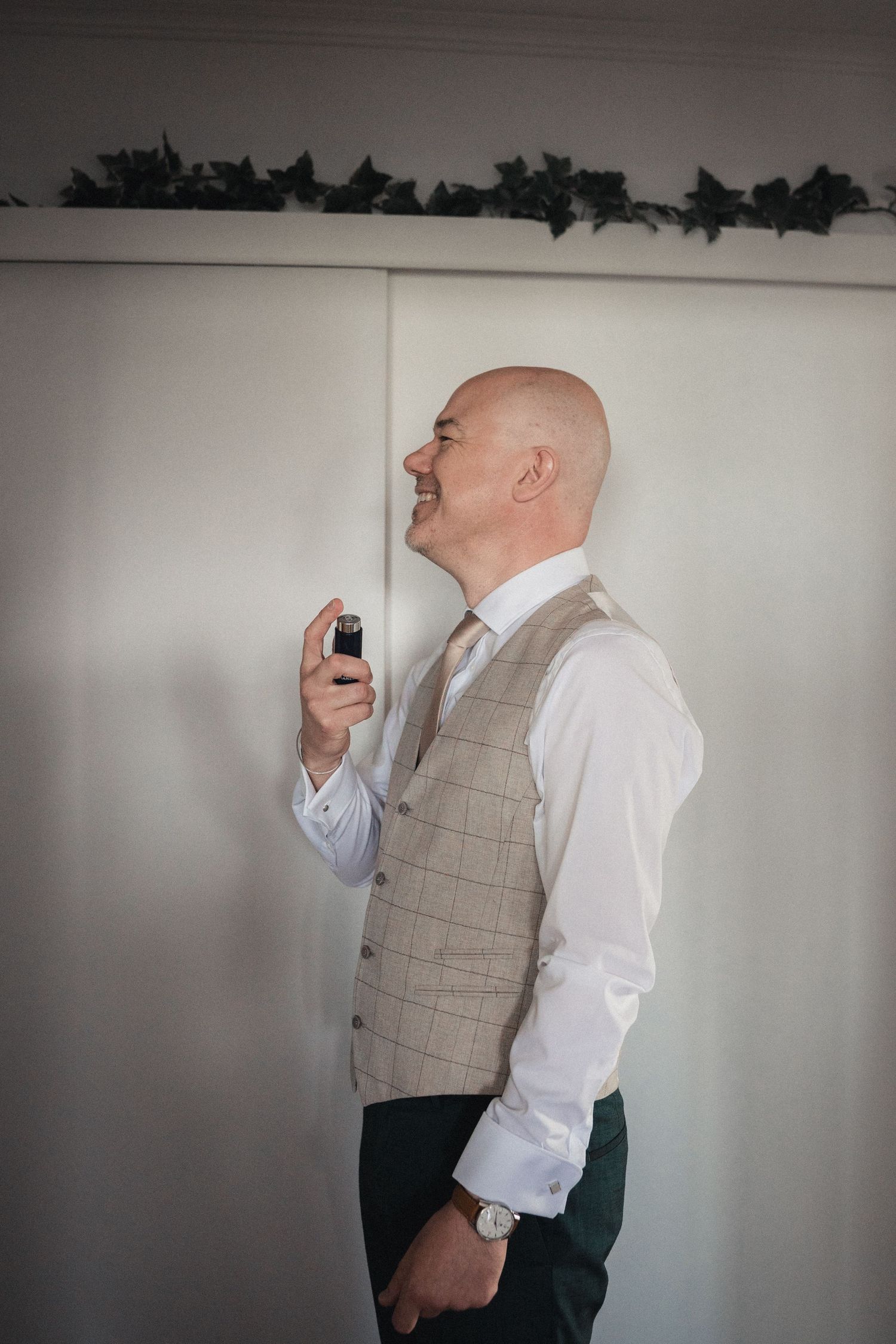 Smiling bald groom in tweed vest and white shirt applying cologne while getting ready for his wedding day.
