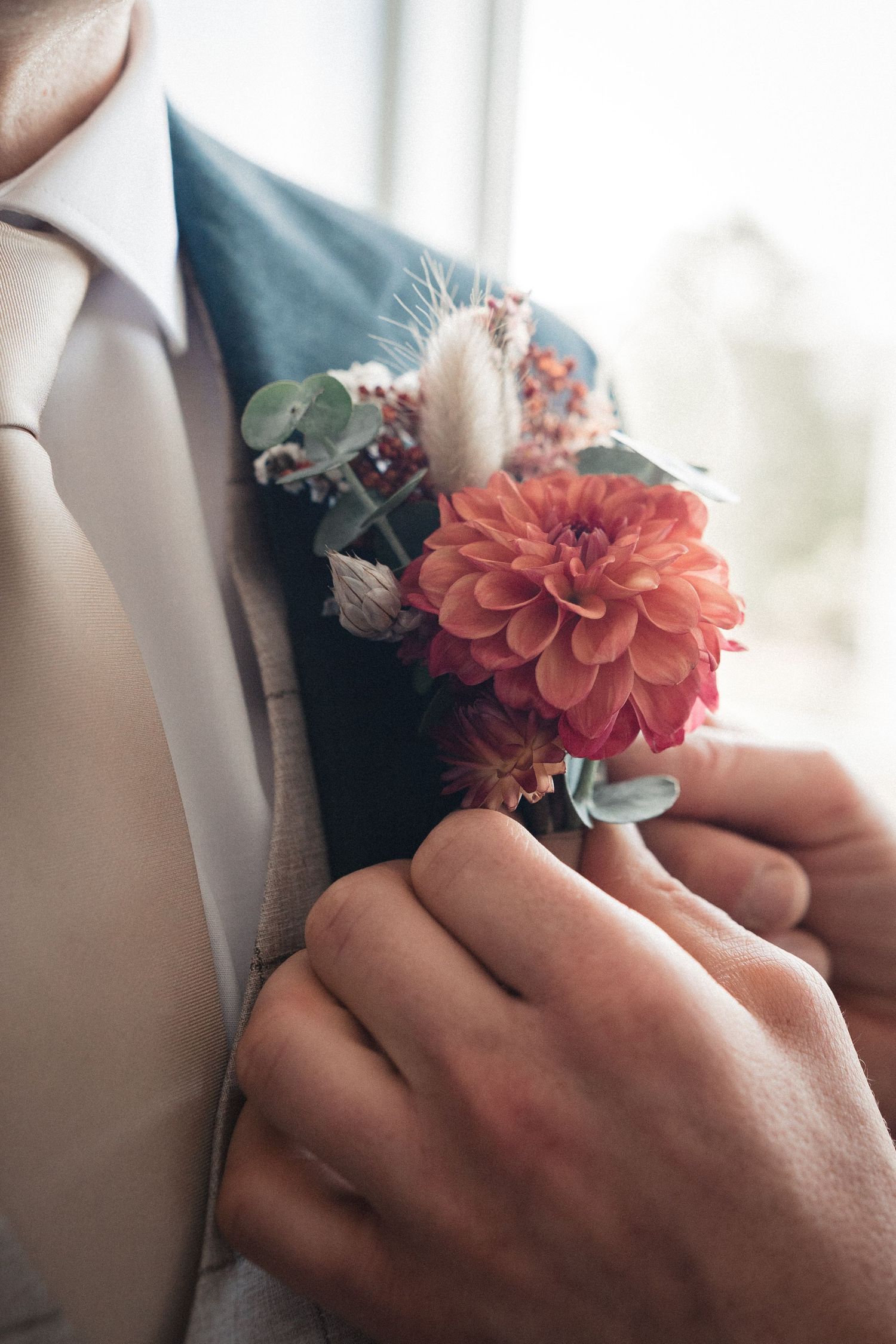 Hand adjusting a coral dahlia boutonniere on a groom's blue suit lapel at a wedding.