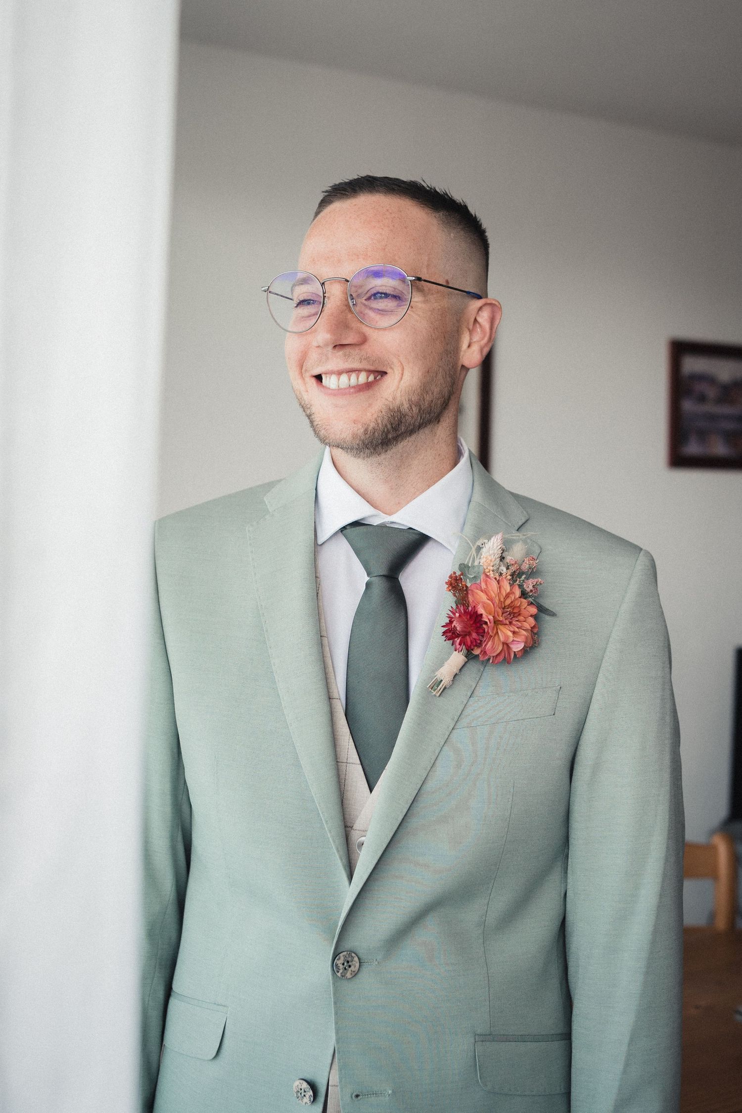 Smiling groom in mint green suit with floral boutonniere and matching tie, wearing round glasses at wedding.