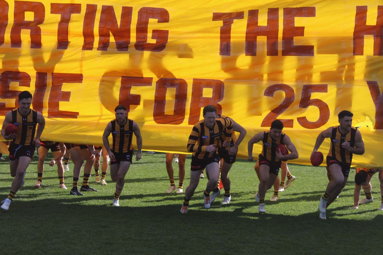Australian football players burst through a gold and brown banner on a green field.