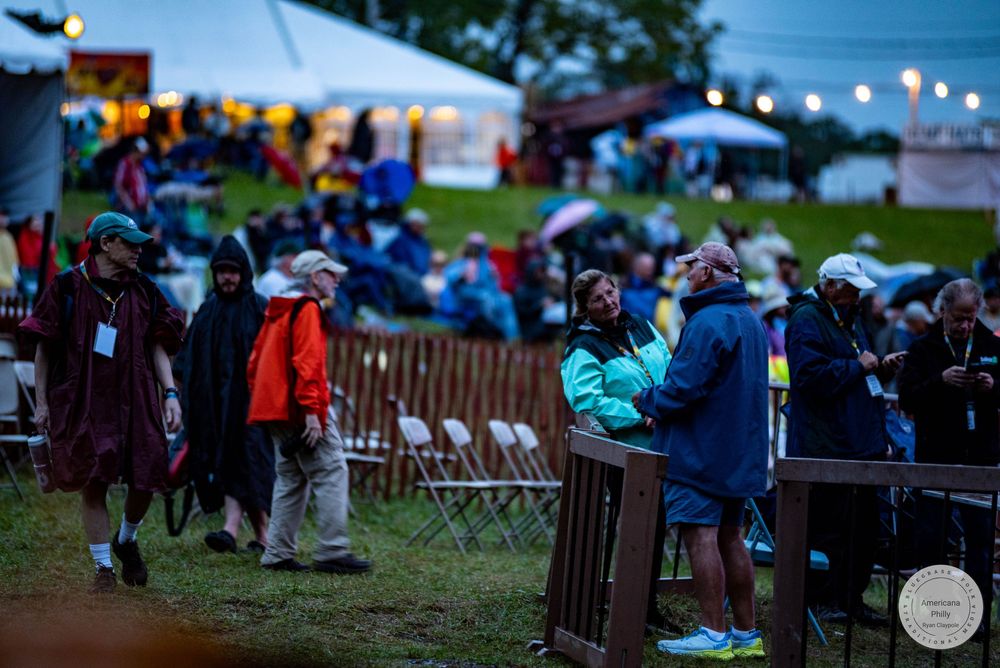 Crowd of festival-goers in rain gear gathered at an outdoor evening event with string lights and white tents.