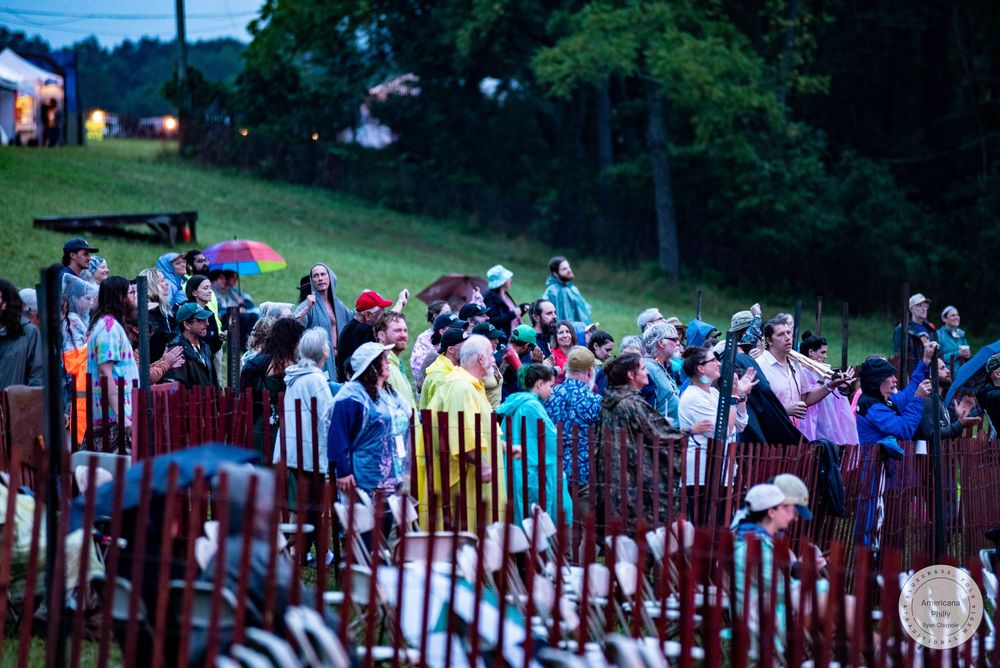 Crowd of spectators watching an outdoor evening event on a hillside, some holding colorful umbrellas in light rain.