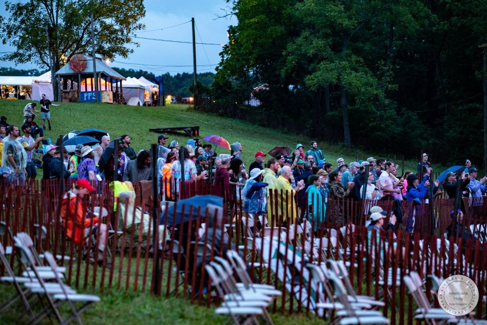 Crowd of people with umbrellas gathered at an outdoor evening festival event on a green hillside.