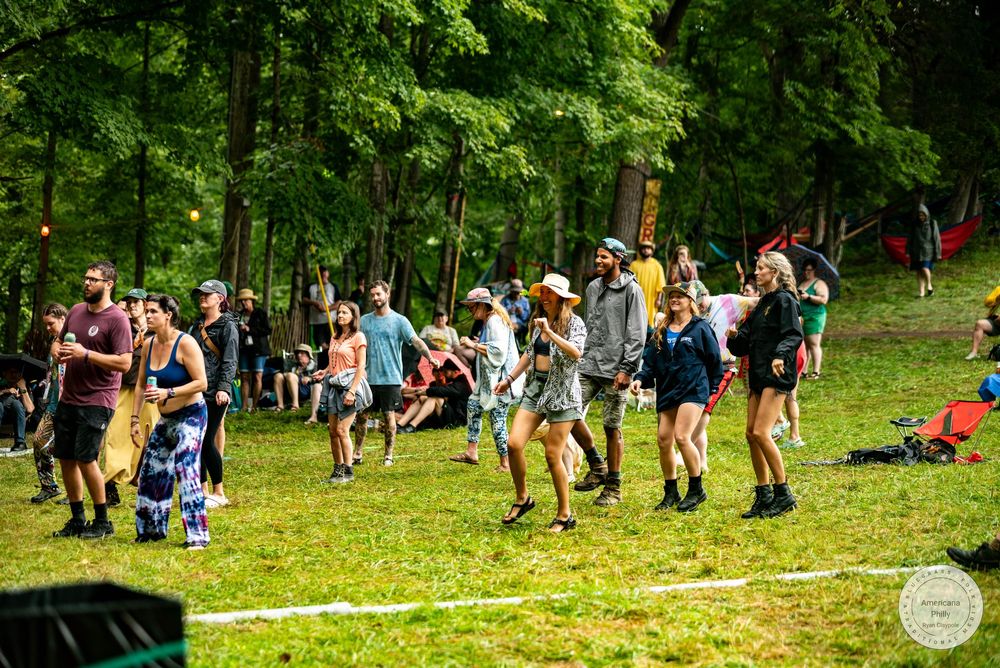 People dancing and socializing outdoors at a summer music festival surrounded by lush green trees.