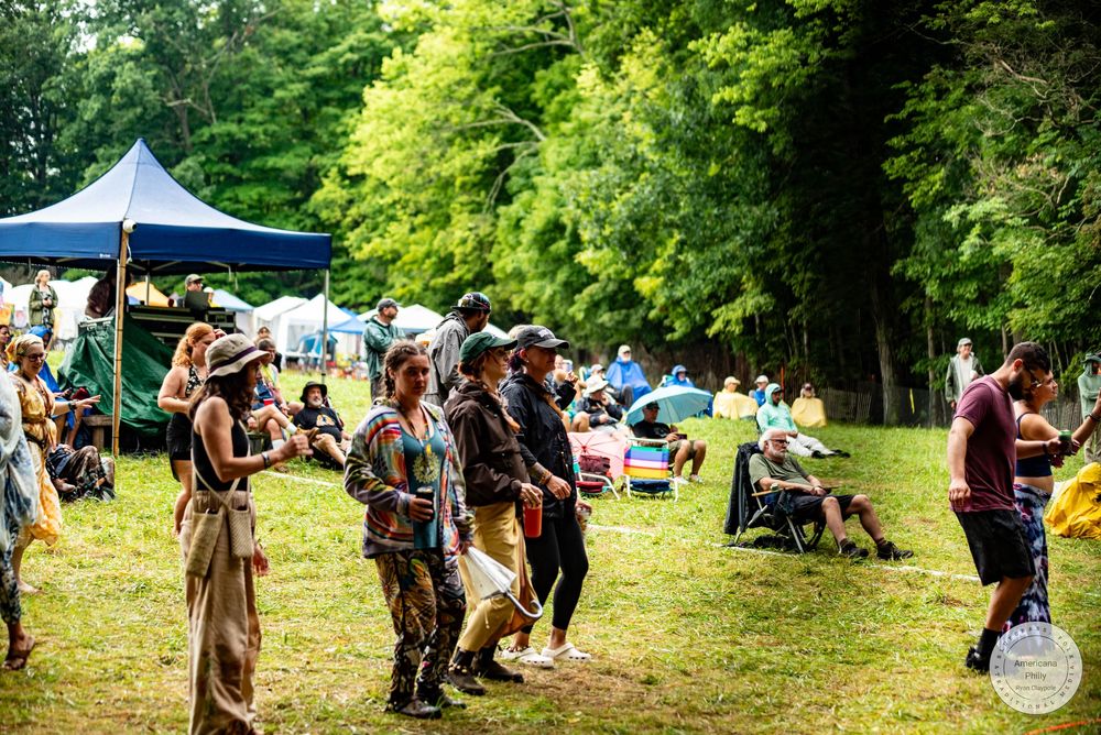 Outdoor festival gathering with people in costumes on a green lawn surrounded by trees and tents on a sunny day.