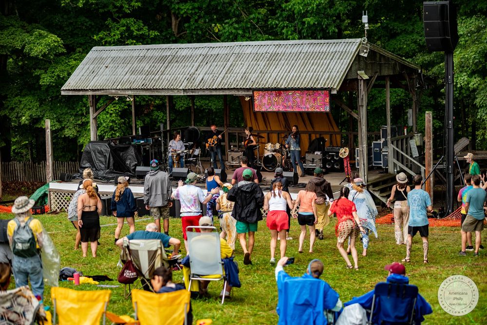 Outdoor music festival with crowd dancing on grass in front of a rustic wooden stage surrounded by lush green trees.