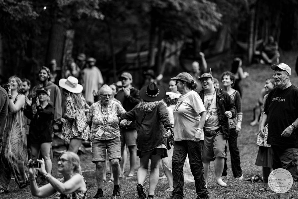 Black and white photo of a crowd of people gathered outdoors at a festival, socializing and dancing among trees.