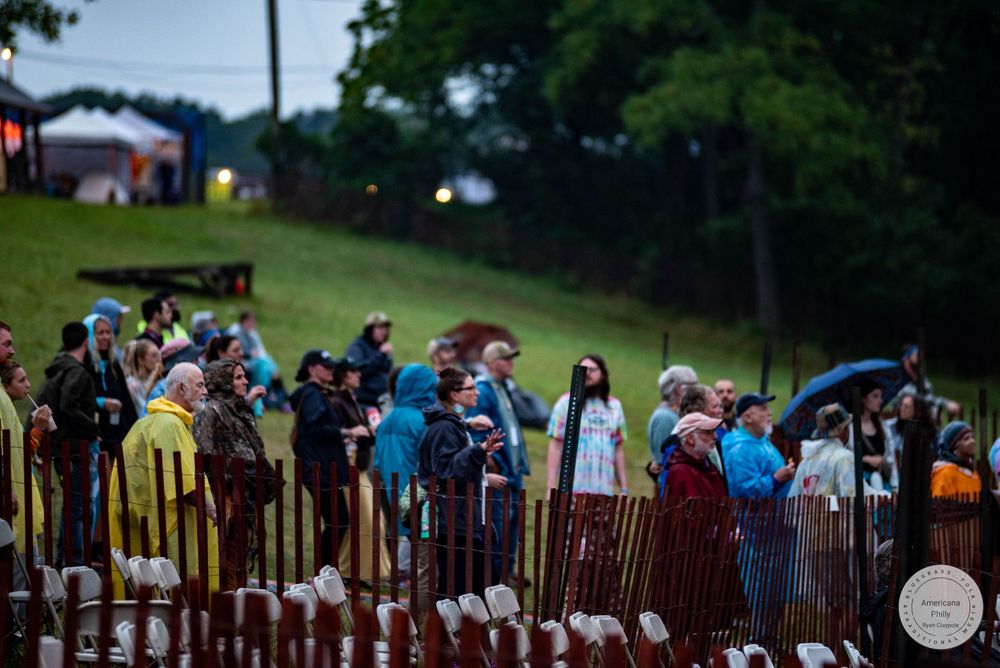 Crowd of festival-goers in rain ponchos standing behind metal barriers at an outdoor music event at dusk.