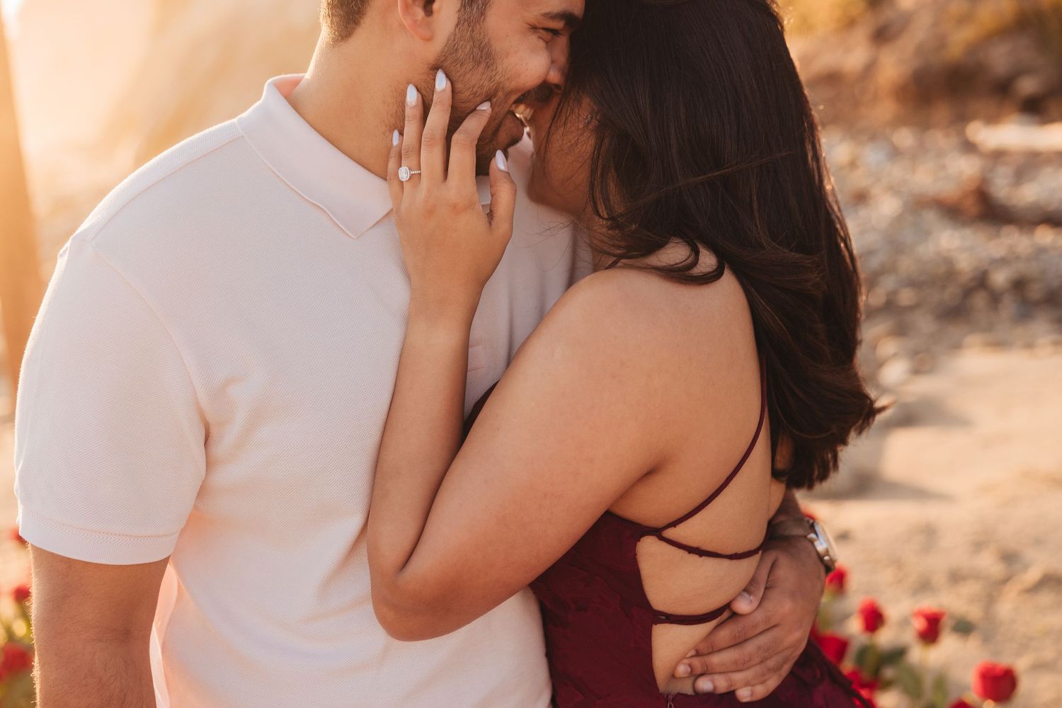 Romantic couple sharing an intimate moment at golden hour, woman in burgundy dress with engagement ring visible.