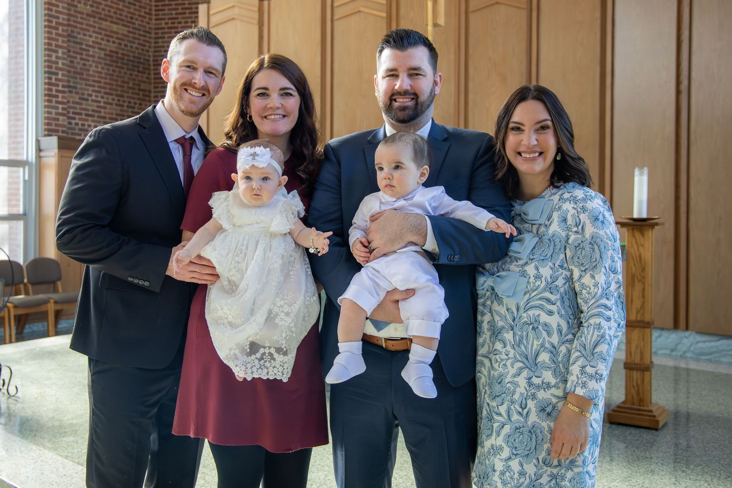 Two couples pose together in a church holding babies dressed in christening and baptism attire.